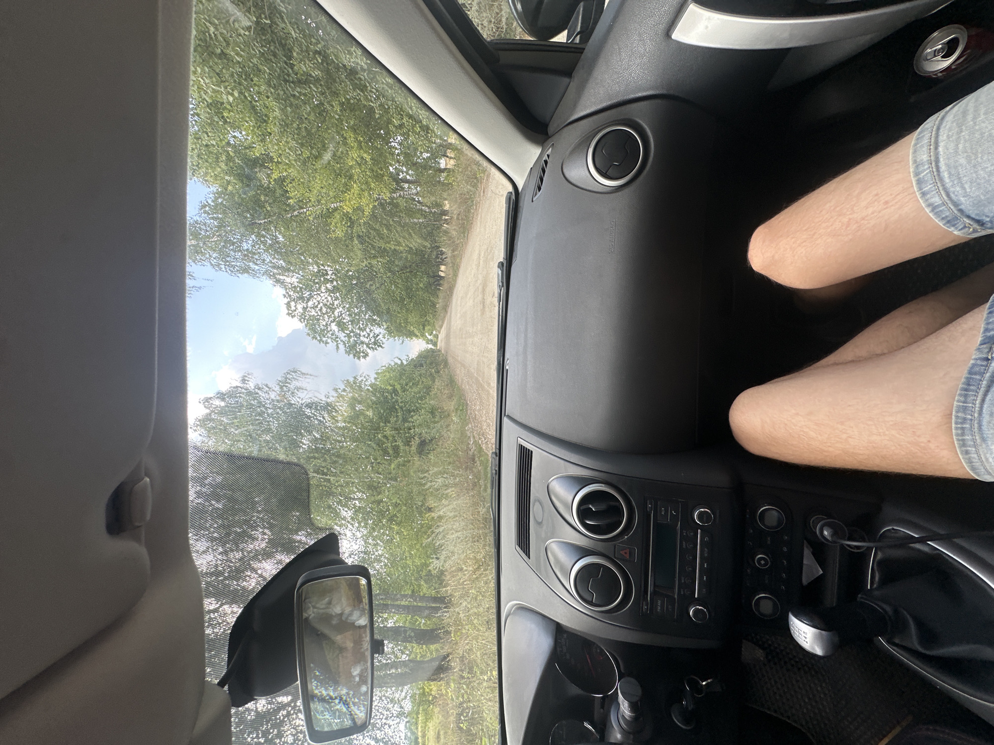 This is a first-person perspective interior shot of a car driving down a dirt road lined with trees. The dashboard is visible, featuring the car's stereo and climate controls. The driver's legs, clad in denim shorts, are positioned in the foreground. The view outside the windshield shows a rural landscape with a dirt road leading into the distance, flanked by dense green trees and a cloudy sky. There is no indication of the city of Tynna, Ukraine in the image.