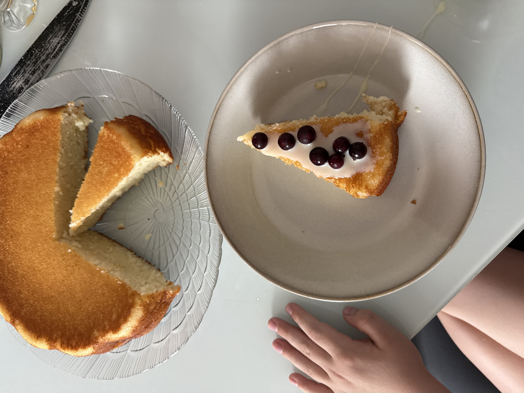 A top-down shot shows two plates of cake on a light-colored surface. The plate on the left is clear with a circular pattern and contains a cake with two slices removed. A slice of cake, topped with white icing and several blueberries, is on the plate on the right. A hand rests near the plate on the right, and part of someone's leg is visible. A knife is visible at the top-left corner. Zhigalki, Russia, is not discernible in this image.