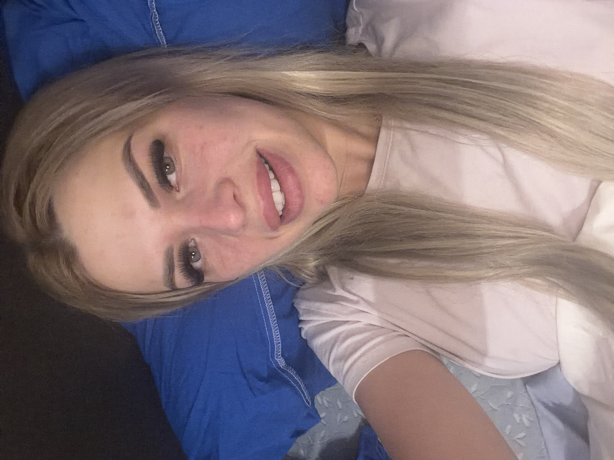 This is a close-up, over-the-shoulder selfie taken by a blonde woman lying down. She is wearing a light pink t-shirt and is positioned against a blue pillow. The background appears to be a bedroom setting, suggesting an indoor environment in Kyiv, Ukraine. The woman is looking towards the camera, with her mouth slightly open, revealing her teeth. Her hair is long and blonde, falling around her shoulders. The lighting in the image is dim, suggesting it might be nighttime or a dimly lit room.