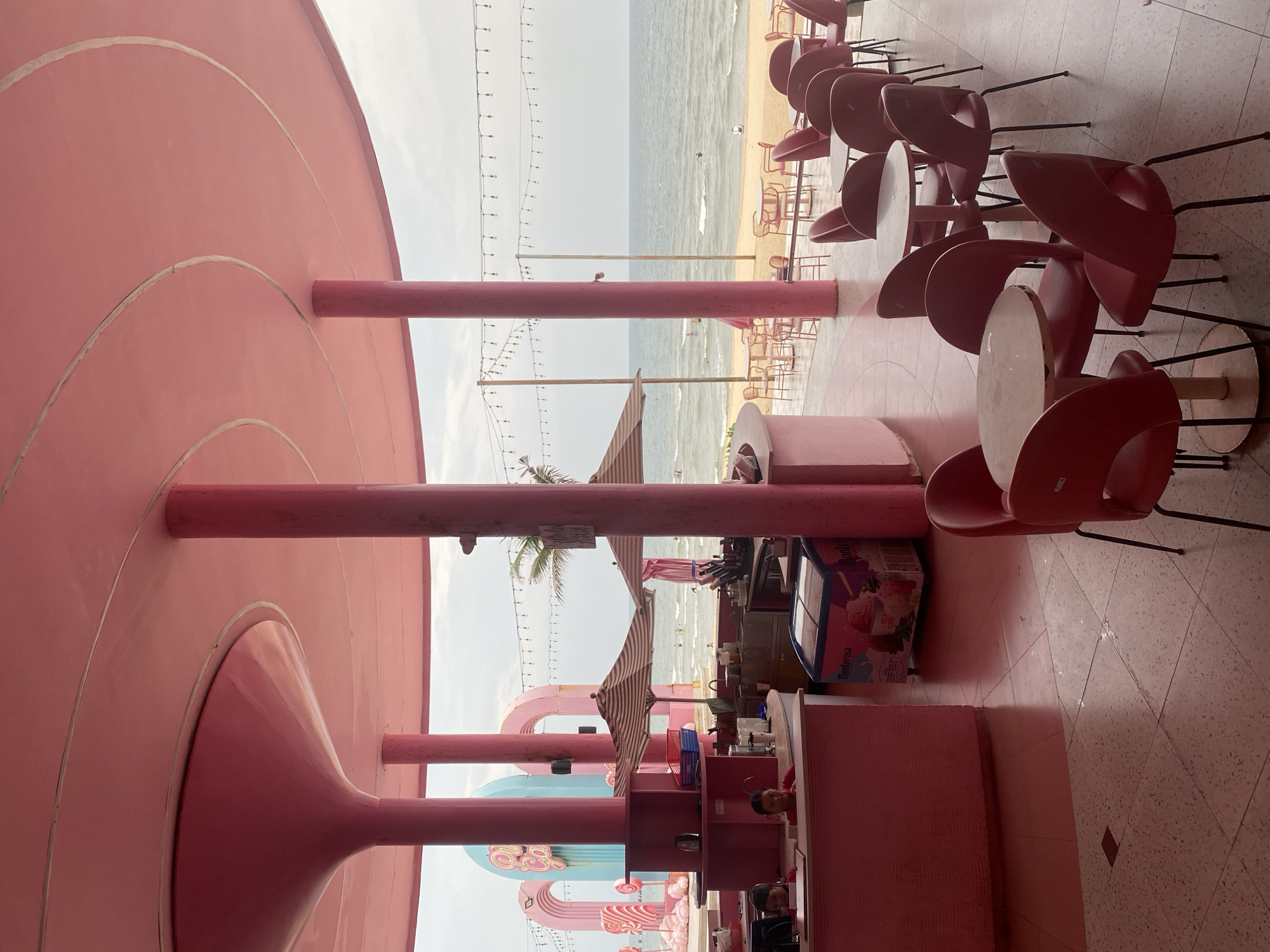 This media file captures a vibrant, pink-themed beachside cafe or restaurant, presumably in Tien Thanh Ward, Vietnam. The image is oriented vertically, showing the establishment's interior and its view of the ocean.
In the foreground, a large, circular pink canopy or ceiling structure features concentric rings, supported by thick, cylindrical pink pillars. Beneath this structure, a seating area is visible with numerous red, modern-style chairs arranged around light-colored, round tables on a tiled floor.
Further back, a counter area includes an ice cream freezer with the brand name "Montarosa" clearly visible on its side. A person is partially seen behind the counter, possibly an employee. Beyond this, playful pink and light blue archways and pillars extend towards the beach, contributing to a whimsical aesthetic. String lights are strung between poles, suggesting an ambiance for evening as well as daytime. A striped beach umbrella and a palm tree add to the tropical setting.
The background reveals a sandy beach leading to a calm, grey-blue ocean under an overcast or hazy sky. Small figures of people can be seen in the water, indicating swimming or wading activities. The overall scene suggests a relaxed and leisure-focused environment, likely a popular spot for visitors to enjoy food and drinks with a beach view during the daytime.