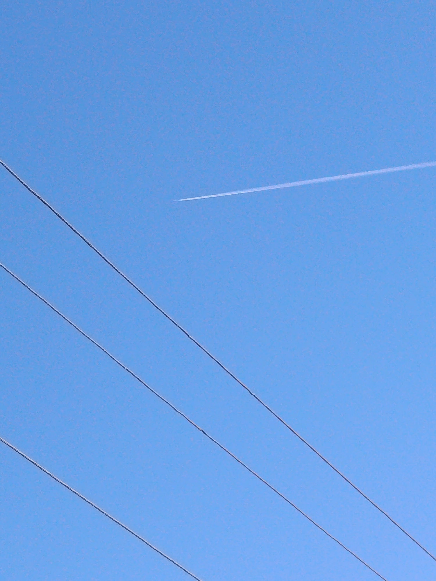 The image shows a clear blue sky with a vapor trail from a jet airplane at the upper right. Four electrical power lines run diagonally from the lower left to the upper right of the frame. I am unable to provide the exact location in the city of Фоки, Russia.