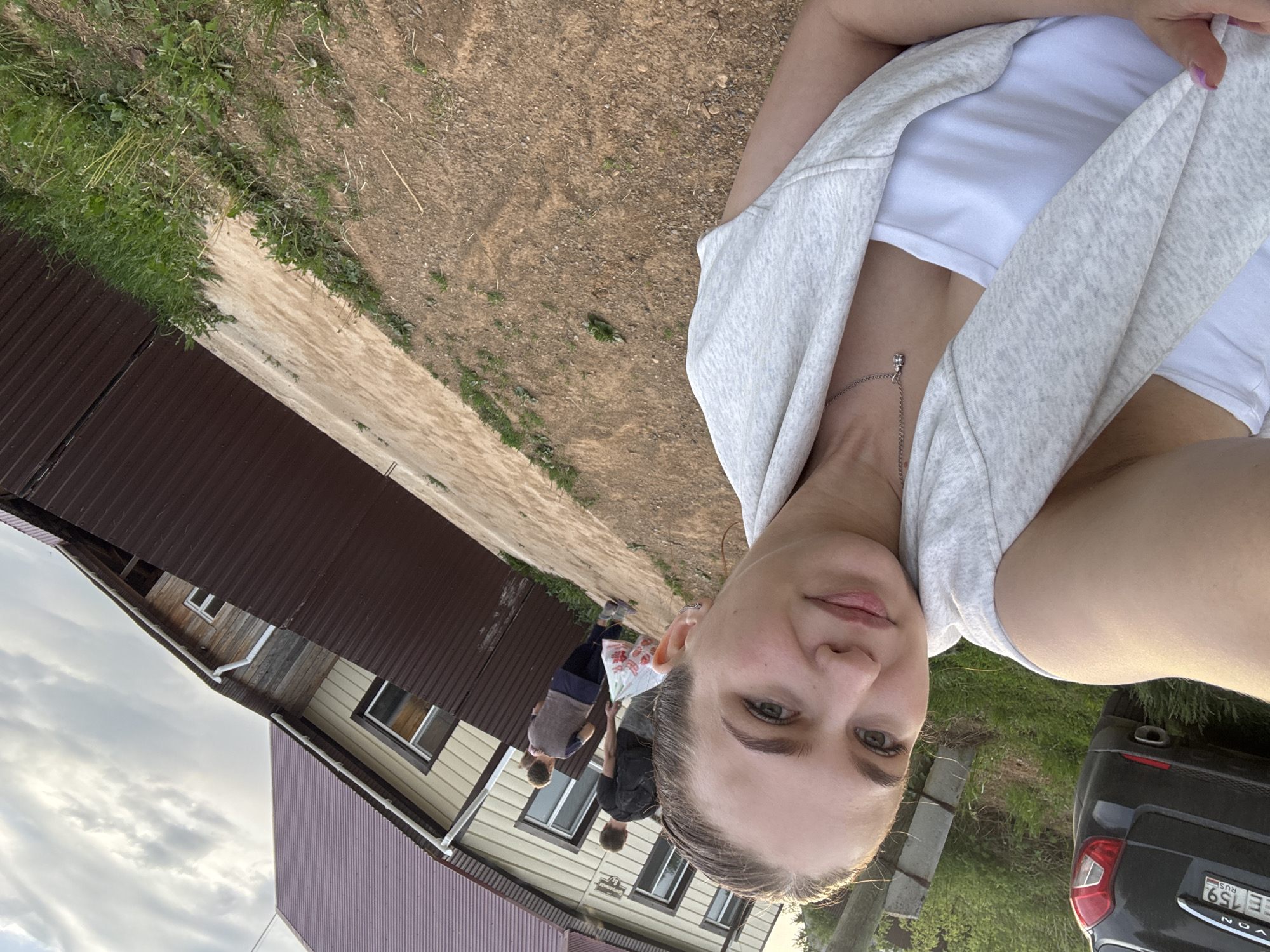 The image shows a selfie taken outdoors. A woman is in the foreground, wearing a white t-shirt and a light grey cardigan. She has light skin and hair, and is wearing a delicate necklace. Behind her, there is a dirt path and a building with a brown metal roof and tan siding. There are three people walking along the path near the building. To the right of the woman is a portion of a dark grey car, with a license plate visible that reads "EE 159 RUS". Based on the information available, the image was taken in Nizhnyaya Gar', Russia. The environment appears to be rural or suburban, with simple buildings and unpaved roads. The lighting indicates it is daytime.