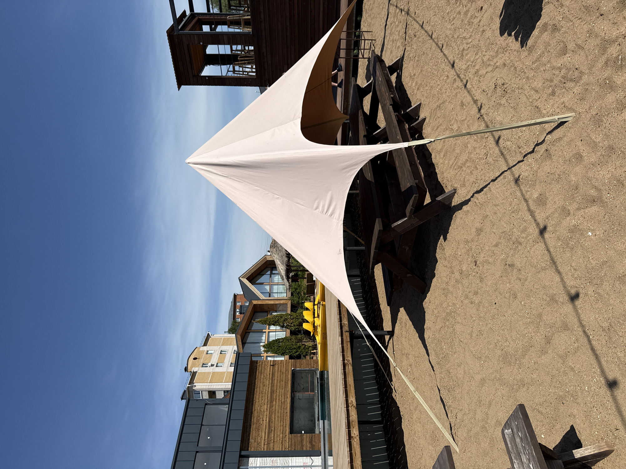 The image shows a sunshade structure adjacent to a picnic table on a sandy surface. The sunshade is a triangular sheet supported by poles. The picnic table is made of dark wood and has attached benches. In the background, there are buildings with mixed architectural styles. There are residential and commercial buildings, possibly in Votkinsk, Russia. The sky is clear and blue. The image is taken in bright daylight.