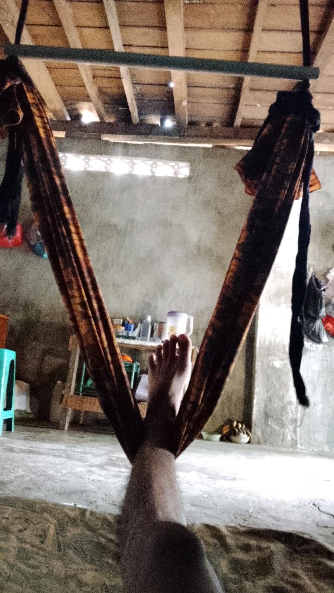 The image depicts an indoor scene, likely a simple dwelling, in Bojong Barat, Indonesia. The perspective is from someone lying down, with their legs extended upwards and partially obscuring the view. They are lying in a hammock made of patterned fabric, which is suspended from a dark horizontal bar attached to a wooden ceiling. The ceiling is constructed with visible wooden beams and planks, with some gaps allowing light to filter through.
The walls of the room are rough and concrete. To the left, a light turquoise object, possibly a stool or chair, is visible. Behind the hammock, a shelf is present, holding various items including bottles and a mug. A wooden structure, potentially a table or support for the shelf, is situated behind the hammock.
In the background, a window with a lattice pattern is letting in light. To the right, dark fabric, possibly clothing, is hanging down. The floor appears to be concrete and is covered in some areas with a patterned fabric or mat. The overall impression is of a humble, everyday living space. The subject's legs are the primary human element in the foreground, indicating a moment of rest or relaxation.