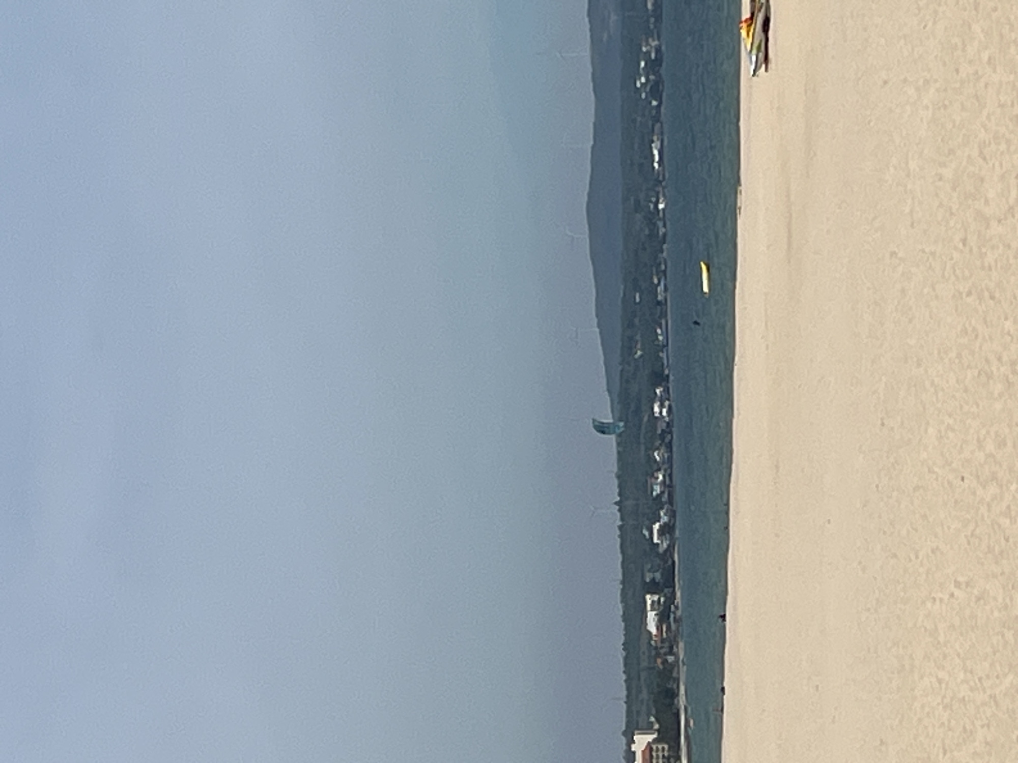 The image depicts a serene beach scene with a calm body of water stretching towards a distant coastline dotted with buildings and hills. In the foreground, a vast expanse of light brown sand occupies the majority of the frame. The sand appears smooth and undisturbed, with subtle variations in tone suggesting gentle undulations.
The middle ground transitions to the water, a tranquil blue-green expanse. A solitary yellow kayak floats on the water's surface. Further in the distance, along the coastline, a cluster of white and beige buildings are visible, nestled amongst lush green foliage. A winding road or pathway appears to connect these structures. Beyond the buildings, rolling hills form the backdrop, topped with several wind turbines, their blades static. A striking blue and curved structure, possibly a sail or a kite, is prominently displayed against the muted grey sky.
The sky itself is overcast, a pale, uniform grey that casts a soft, diffused light over the entire scene. The overall impression is one of peacefulness and tranquility, a typical coastal landscape under a cloudy sky. The location is Phu Thuy Ward, Vietnam.