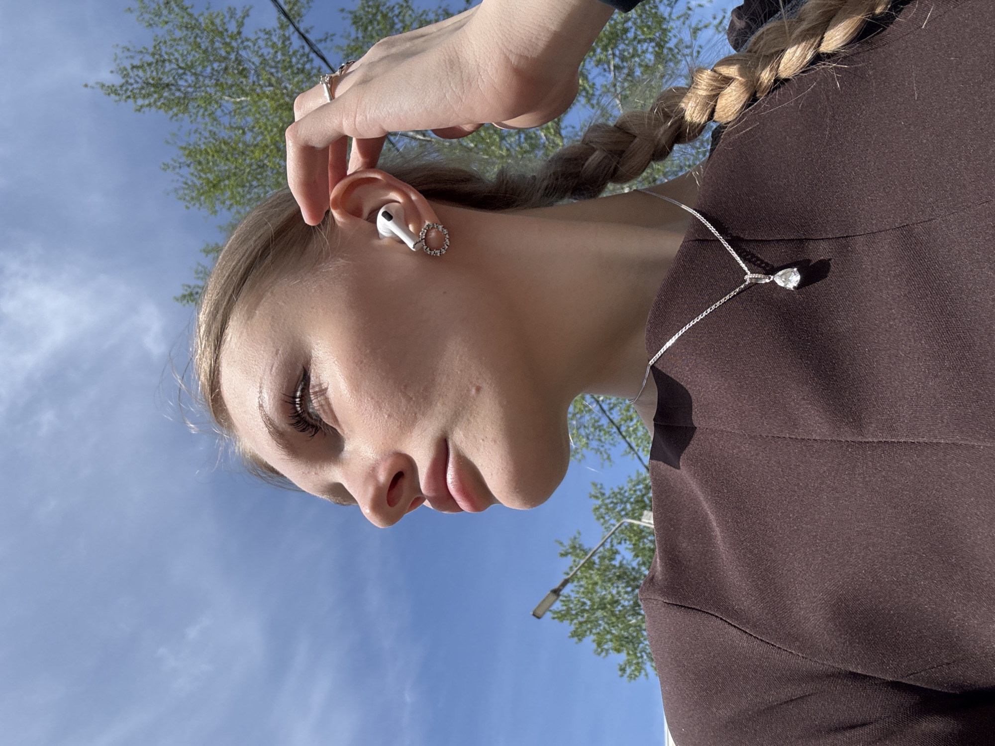 A young, light-skinned woman with blonde hair braided and pulled back, is seen from a low angle against a backdrop of a blue sky and green trees. She is wearing a brown blouse and a silver necklace with a teardrop pendant. She has a white wireless earbud in her ear, adorned with a circular, possibly jeweled, accessory. Her right hand is raised, seemingly adjusting or touching her ear. The location appears to be an outdoor setting, likely in Perm, Russia based on the prompt instructions, with what seems to be a street light pole visible among the trees.