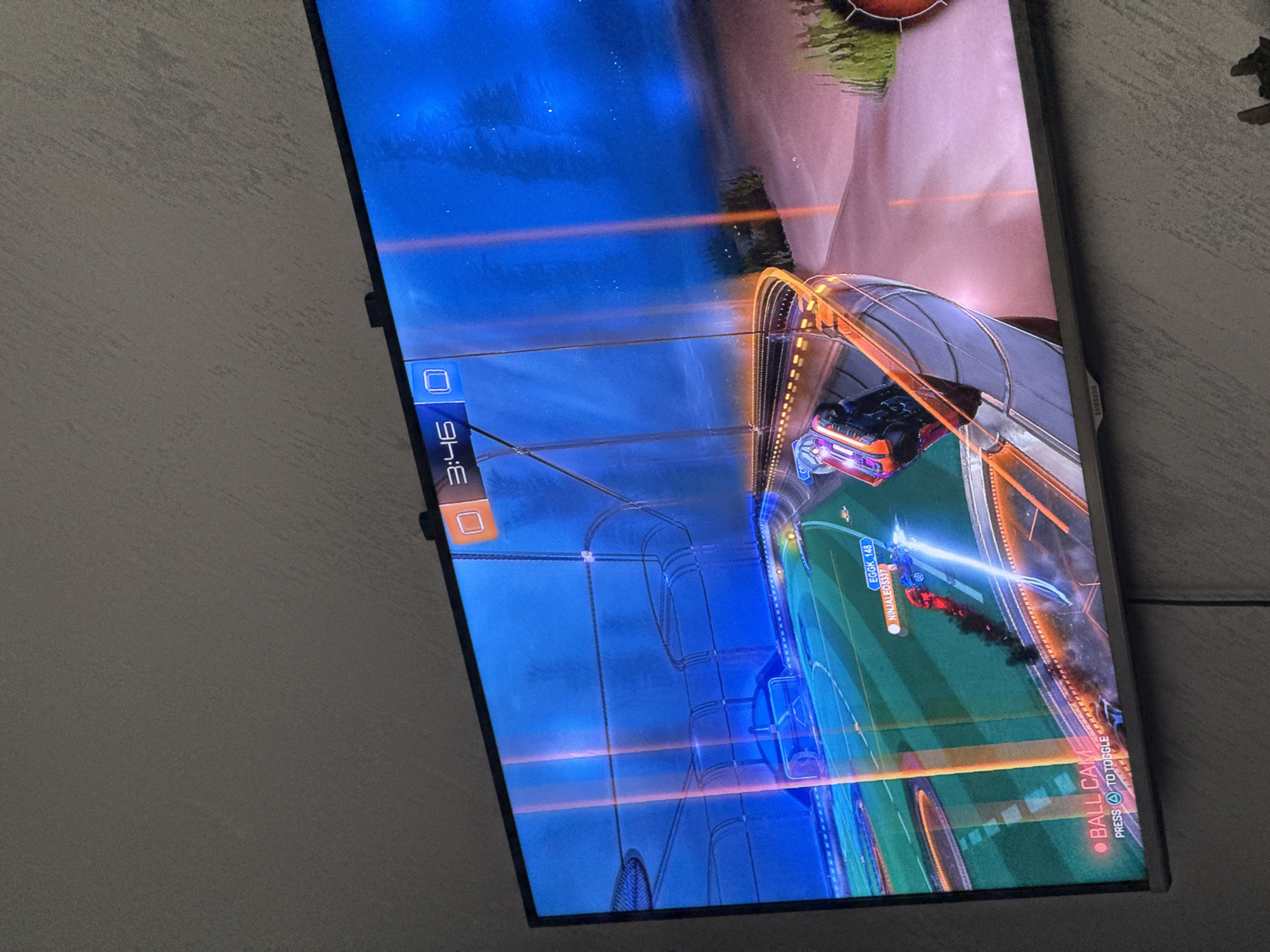 The image depicts a television screen displaying gameplay from the video game "Rocket League". The game shows two cars competing in a soccer-like match in an enclosed arena. The time remaining in the match is 3:46.
The location is not in Glojene, Bulgaria. There is no indication of the location.