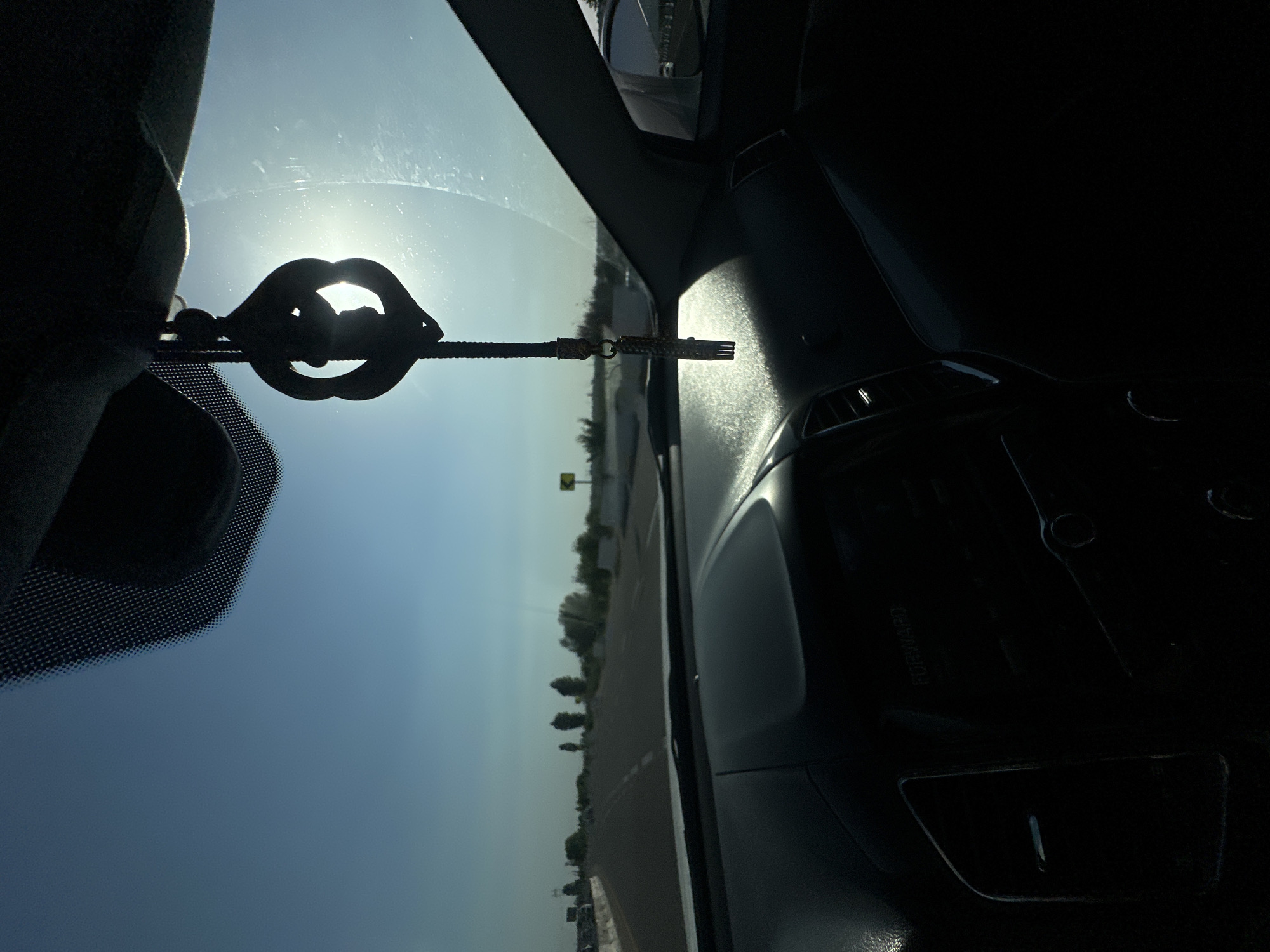 The image is a photograph taken from inside a vehicle, looking out through the windshield. The sun is bright and directly in view, creating a lens flare and obscuring details outside. A car accessory, shaped like lips and attached to a string, hangs from the rearview mirror, silhouetted against the sun. The interior of the car is dark, with a dashboard visible on the right side. The exterior shows a road with trees lining the sides, suggesting an outdoor setting, possibly a highway or a road through a semi-rural area. The sky is a bright blue, indicative of a clear day. The orientation of the image is sideways. There is no discernible text. The scene appears to be captured during the daytime, likely mid-day due to the intensity of the sun. The location context is Tashtugizak, Uzbekistan, which aligns with the arid-looking landscape and the type of vegetation visible.