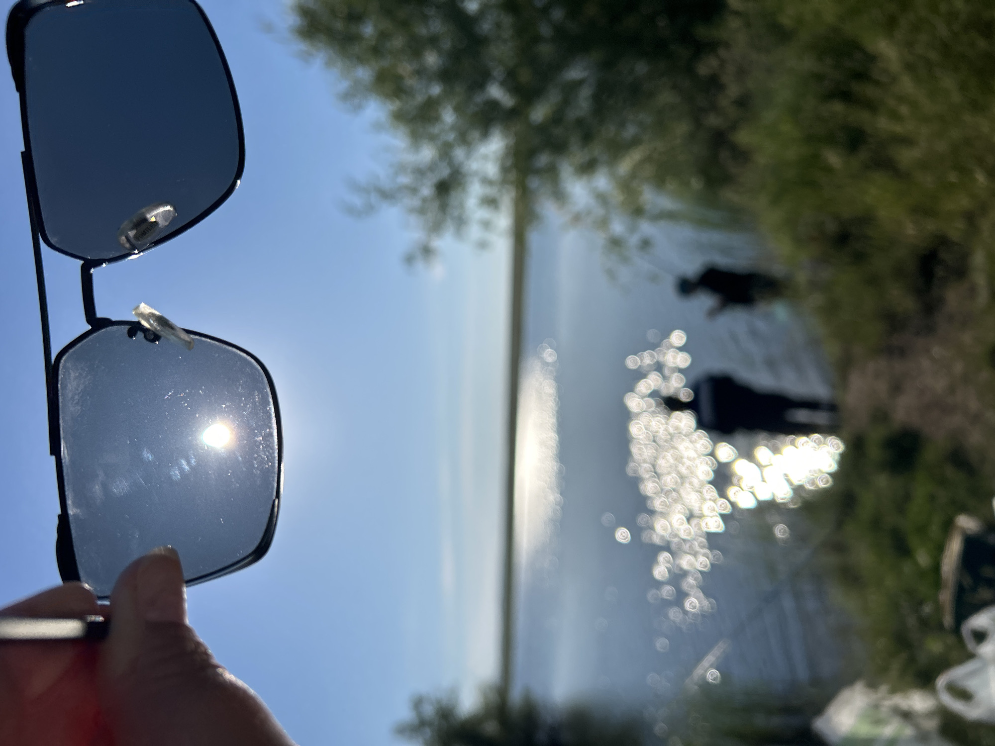 An over-the-shoulder shot captures sunglasses held up by a hand in the foreground, obscuring the upper portion of the image. Through the lenses of the sunglasses, the sun is visible against a clear blue sky. The scene beyond the sunglasses depicts a body of water, likely a lake or river, reflecting the bright sunlight with numerous bokeh highlights. Two silhouetted figures are standing near the water's edge, one closer to the viewer than the other, both appearing to be fishing. The background is softly blurred, with green vegetation visible on the right side of the frame and along the shoreline. The image is taken outdoors, under bright daylight conditions. No specific location in Matrayevo, Russia is identifiable from the visual information in the image.
