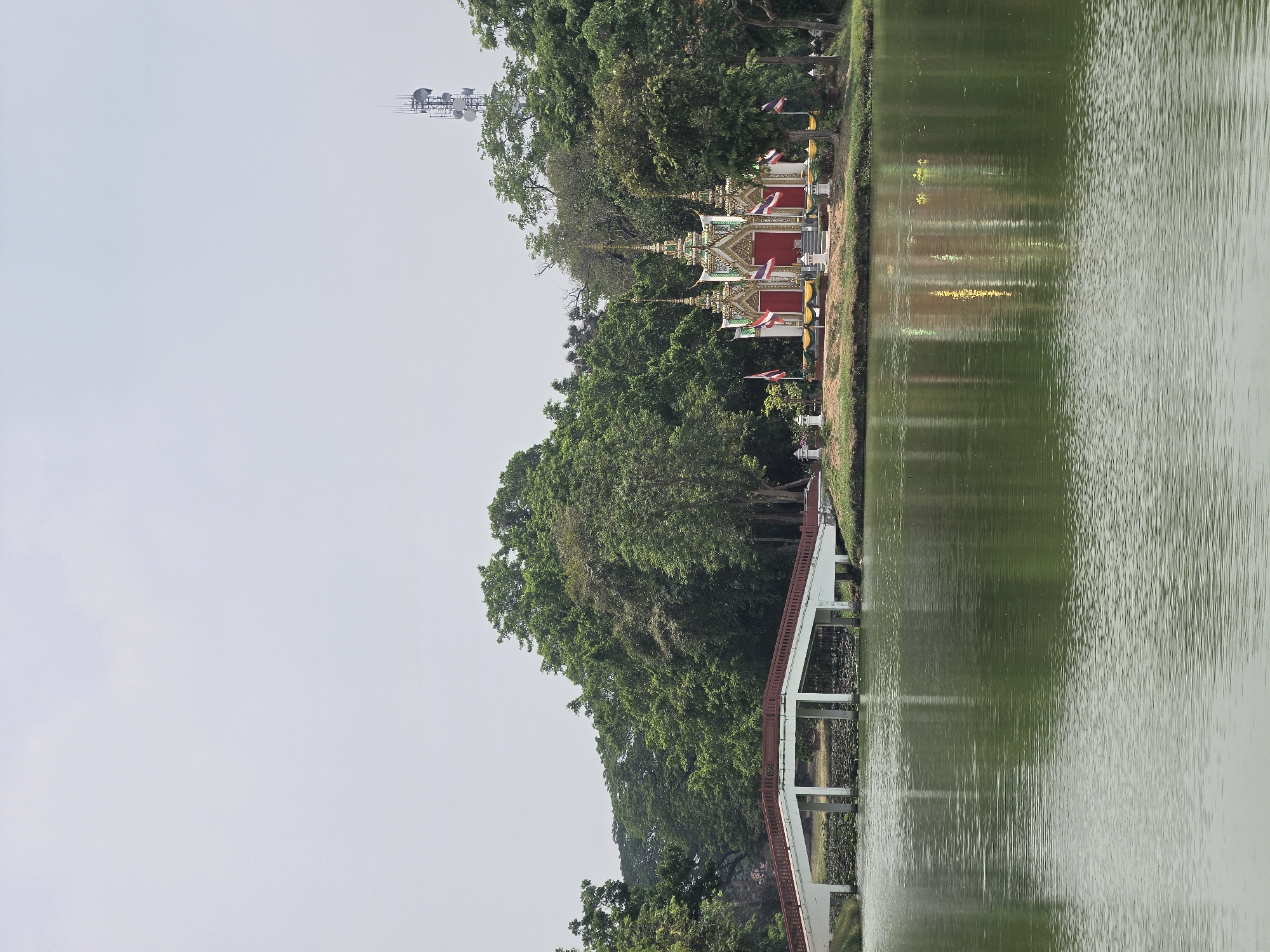 This image depicts a serene waterside landscape in Phra Nakhon Si Ayutthaya City Municipality, Thailand. The right side of the frame is dominated by a calm, greenish-brown body of water, likely a lake or canal, displaying gentle ripples and subtle reflections.
Along the left bank, a lush expanse of green trees creates a dense, verdant backdrop. Nestled among the foliage are several small, ornate traditional Thai shrines or pavilions. These structures are characterized by their red walls, white bases, and multi-tiered roofs detailed with golden and green accents, reflecting typical Thai architectural styles. Small Thai flags, featuring red, white, and blue stripes, as well as some yellow and white flags, are visible near these shrines.
Further along the bank, a white bridge or walkway with reddish-brown railings extends, appearing to span or run alongside the water. Above the treeline in the upper-left, a modern communication tower equipped with multiple antennas stands out against the light, overcast sky, providing a contrast to the traditional and natural elements.
The soft, diffused lighting suggests an overcast day or a time of day without harsh sunlight. The scene is devoid of people, contributing to its tranquil and peaceful ambiance. No discernible text is visible in the image.