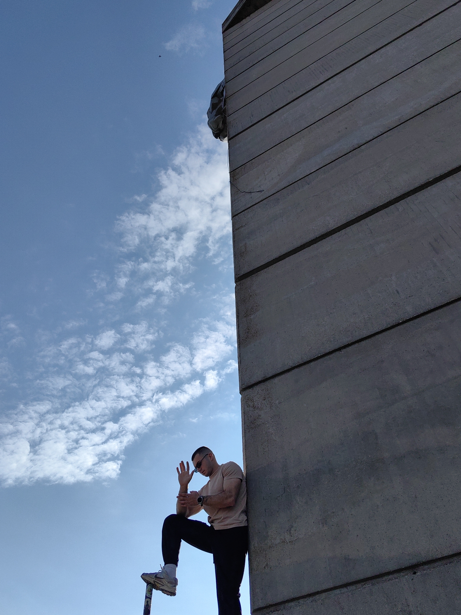A low-angle shot depicts a man standing next to a tall concrete structure under a partly cloudy sky. The man is wearing a light brown t-shirt, dark pants, sunglasses, and sneakers. His right leg is raised, with his foot resting on a narrow vertical object near the bottom of the frame. He is facing towards the left side of the frame and has his right hand raised, fingers splayed. He is also wearing a watch on his left wrist.
The concrete structure is composed of horizontal slabs and takes up a significant portion of the right side of the frame. It appears to be part of the Founders of the Bulgarian State Monument in Shumen, Bulgaria.
The sky is blue with scattered white clouds. A small, dark object, possibly a bird, can be seen in the upper center of the frame.