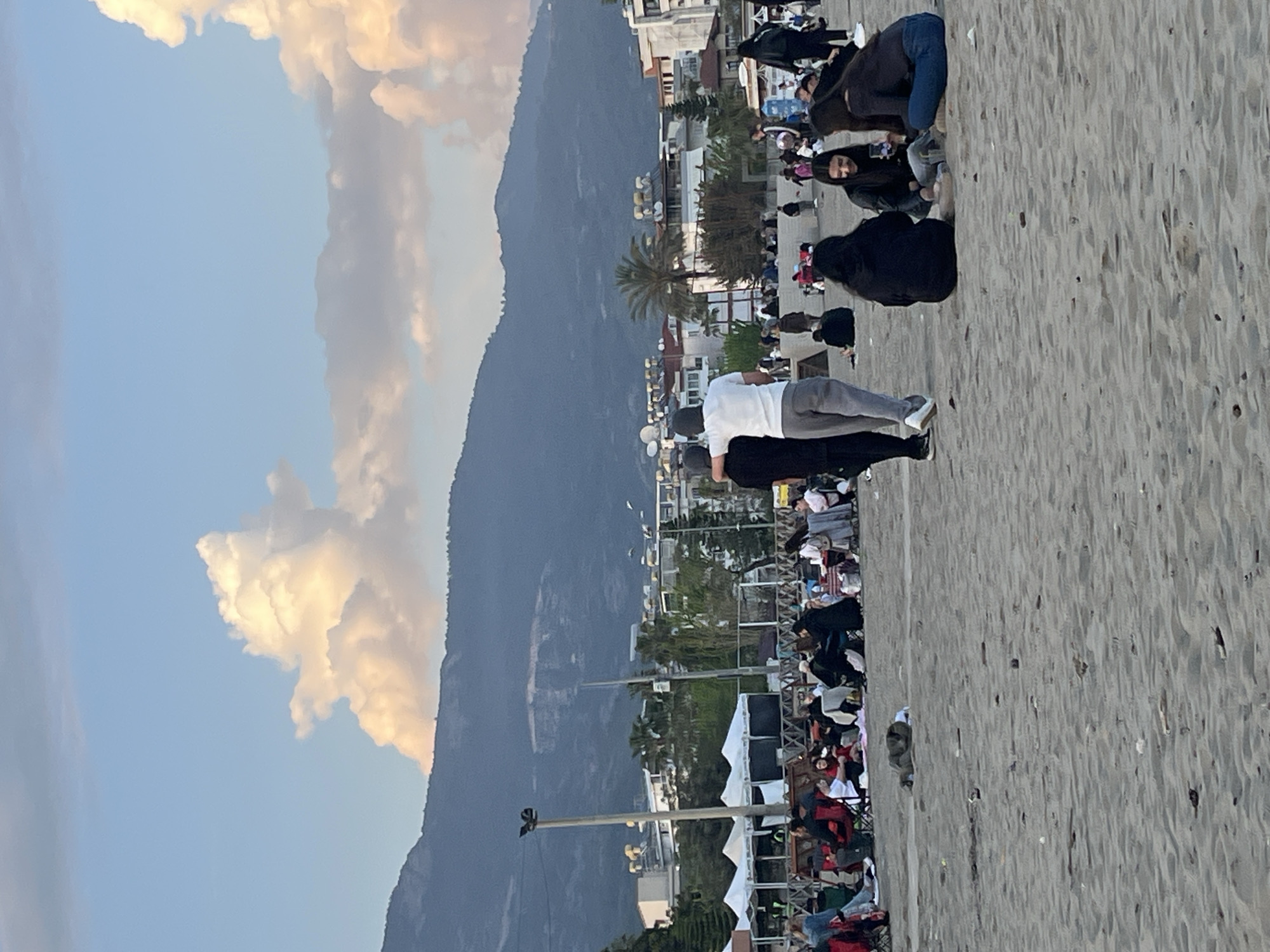 The image depicts a crowd gathered on a beach in Alanya, Turkey. In the background are buildings and a mountain range under a cloudy sky. People are standing and sitting on the sand, some appearing to be conversing. A series of white tents are set up, possibly for an event. The immediate foreground is the sandy beach.