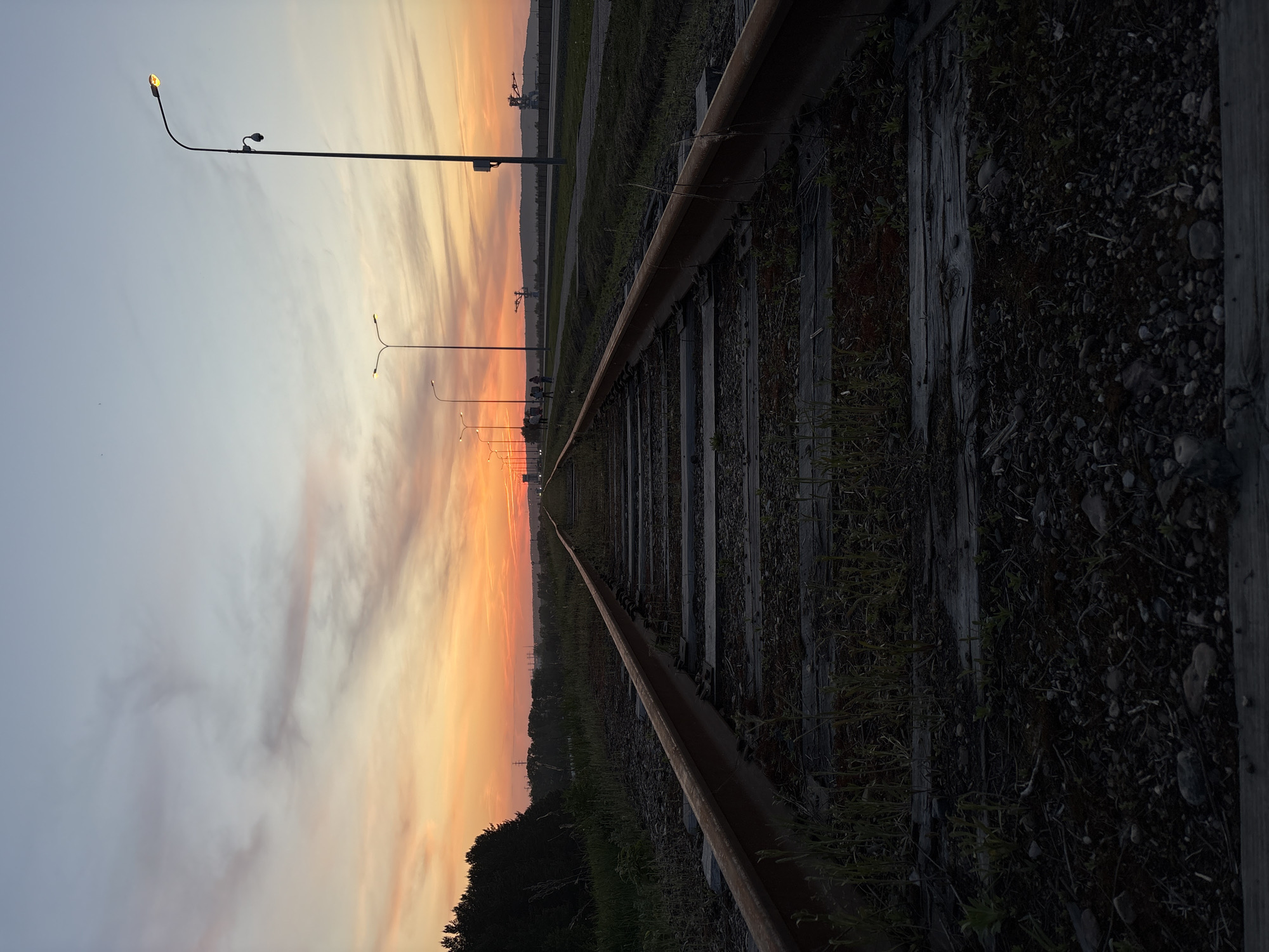 The image depicts a railway track stretching into the distance under a sunset sky. The sky transitions from a pale blue-grey at the top to a vibrant orange and red near the horizon. Silhouetted against the sunset are street lights lining the path along the track. The tracks themselves are rusty and partially obscured by vegetation. Wooden sleepers form the base of the track, visible between the rails. In the far distance, possibly beyond the city limits, are industrial cranes. The image is rotated to the side, so the sky is on the left, and the ground is on the right. This scene is possibly located in the city of Chaikovsky, Russia.