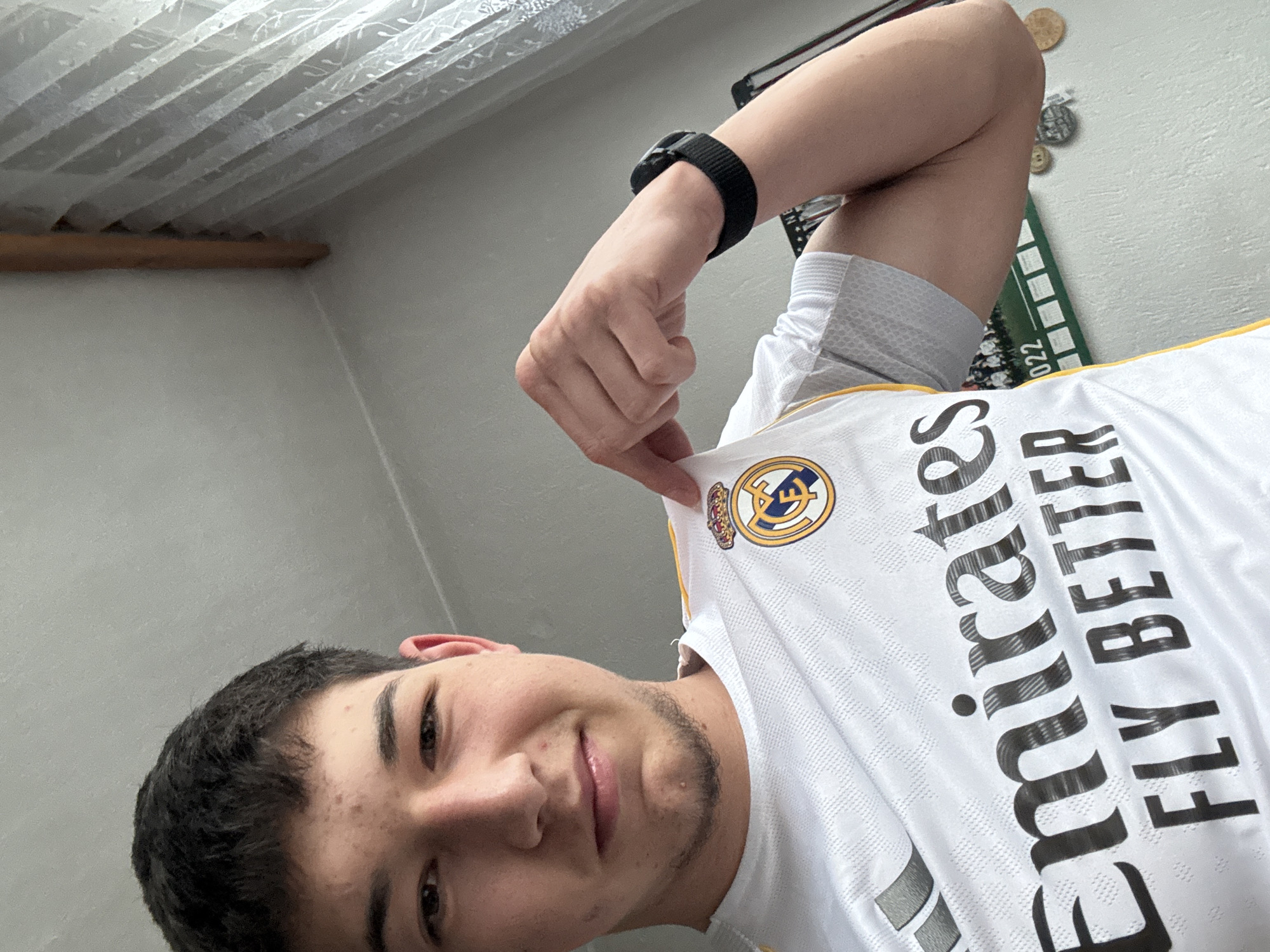 A young man poses for a selfie indoors, likely in Zlatograd, Bulgaria. He is seen from a slightly upward angle, suggesting he might be lying down. He has short, dark hair, dark eyes, and a faint smile. A light mustache and chin beard are visible, along with some blemishes on his forehead. He is wearing a white short-sleeved football (soccer) jersey, identified as a Real Madrid kit by the prominent crest on the upper left chest, which features a golden circle, blue lettering, and a crown. The main sponsor, "Emirates FLY BETTER," is printed in large black letters across the front of the jersey. A black watch or fitness tracker is worn on his left wrist.
The background shows a plain, light-colored wall. In the upper left portion of the image, a window is visible, covered by a white, translucent curtain with a subtle pattern. A wooden curtain rod runs horizontally below the curtain. On the upper right side of the wall, several circular metallic objects, resembling coins or medals, are affixed as decoration. Below these, a green and white rectangular item, possibly a calendar or display board, is partially visible, showing the year "2022." The bright, natural light filtering in suggests the photo was taken during the daytime. The man holds the collar of his jersey up slightly with his right hand, showcasing the emblem.