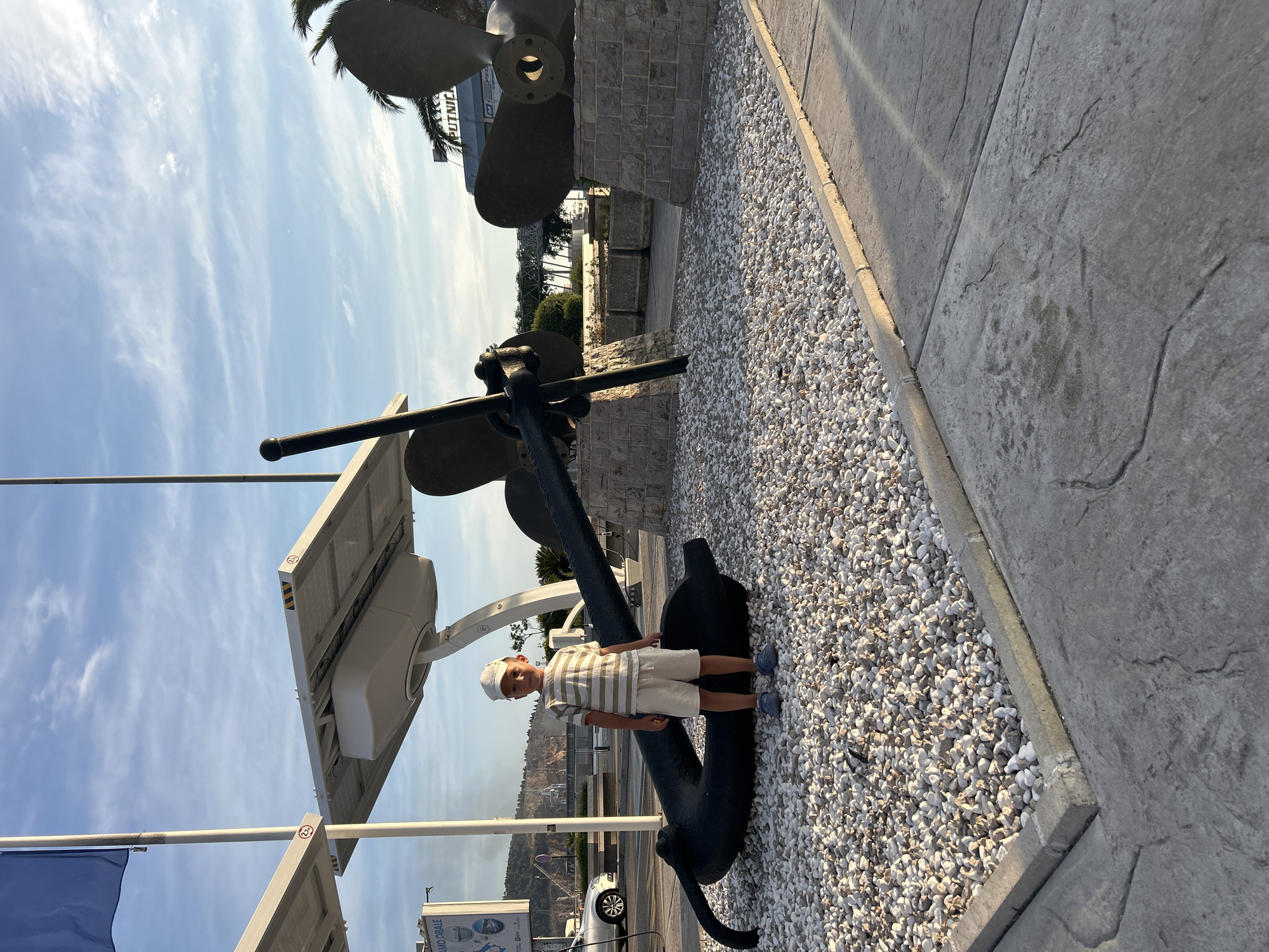 A young boy in a striped shirt, white shorts, and a white hat stands next to a large, dark-colored anchor. He is smiling at the camera. The anchor appears to be a monument, situated on a bed of small white pebbles. Behind the anchor, there is a large ship's propeller mounted vertically on a stone base. The scene is outdoors, under a bright blue sky with scattered white clouds. The sun is casting long shadows, suggesting it is late afternoon or early morning.
In the background, there are palm trees, some buildings, and a body of water with boats. A white car is visible parked on a road to the left, along with a blue flag and a sign with text. One sign partially visible reads "ROBIE QUALE". The overall setting appears to be a waterfront area, possibly a promenade or a park near a marina. The weather is clear and sunny. The boy's expression conveys a sense of happiness and perhaps enjoyment of his surroundings. The large nautical elements suggest a connection to the sea and maritime history.