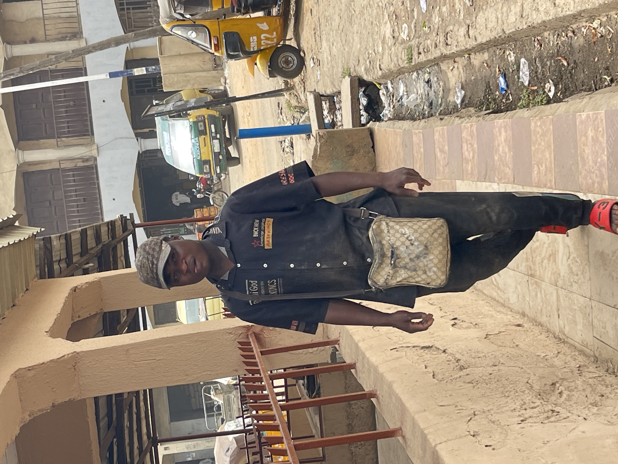 A medium outdoor shot shows a man standing on the sidewalk in Zaria, Nigeria. The man, who appears to be in his 30s, has dark skin and is wearing a black button-down shirt with patches and text, dark-colored pants, a patterned cap, and red sandals. A patterned bag hangs across his shoulder. He is facing and looking towards the camera.
Behind the man, three-wheeled vehicles known as "Keke Napep" are parked along a street. A building with barred windows is visible in the background. A concrete drainage ditch with trash runs alongside the sidewalk. The sidewalk is made of light-colored rectangular tiles. The lower part of the image shows a beige building with rust-colored metal railings and a low wall.