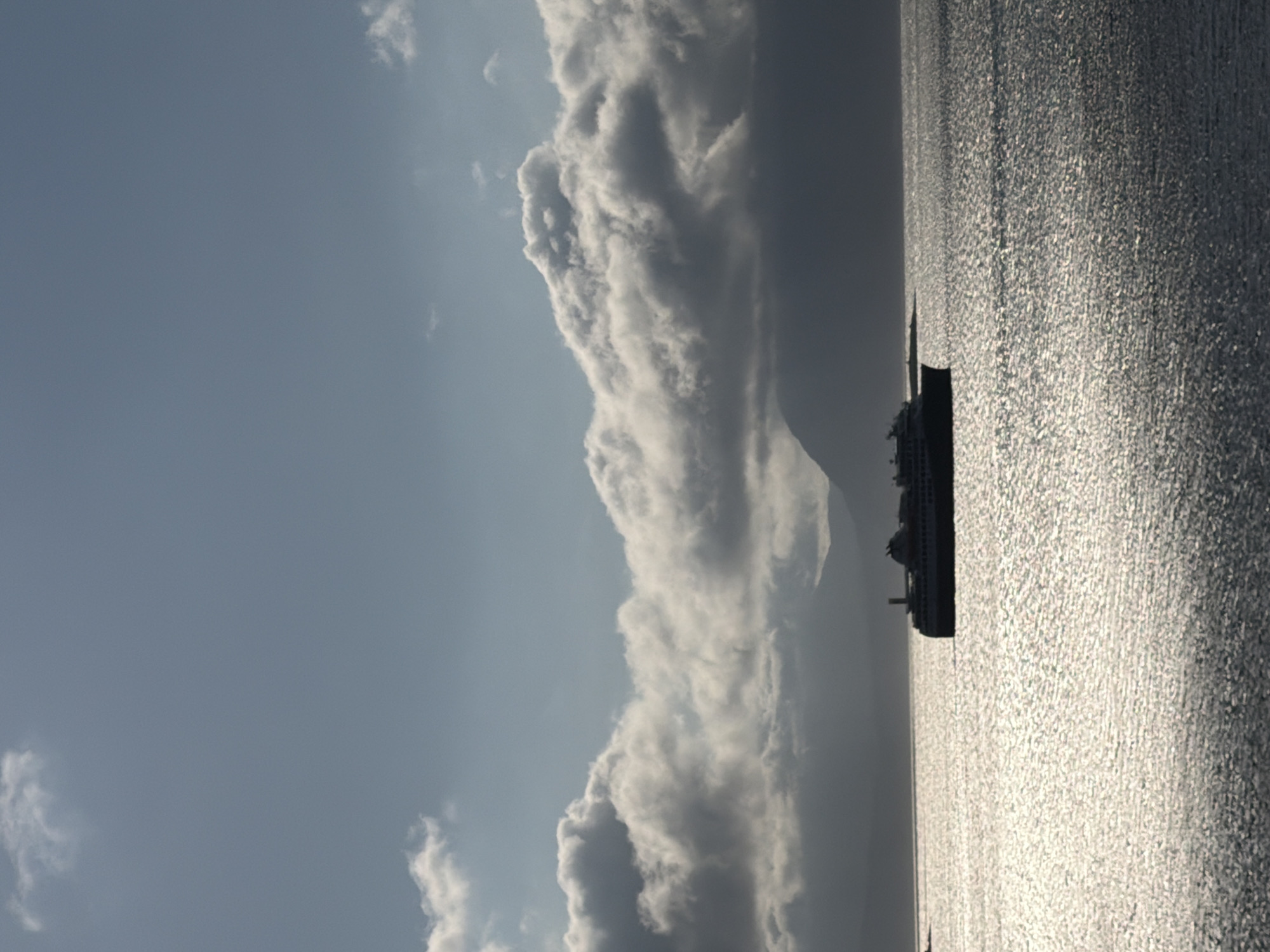 A large ferry is sailing on the choppy, sunlit water in front of a silhouette of mountains under a cloudy sky. The ferry is a dark silhouette against the bright water. The sky is a pale blue with scattered white clouds, some of which are dark and heavy. The water is very reflective, with the sun creating a shimmering effect. The mountains in the background are dark and indistinct, suggesting they are distant. The scene appears to be a calm day at sea, with the ferry the main subject of interest. This scene is likely located in the waters near Harstad, Norway, given the presence of mountains and ferries.