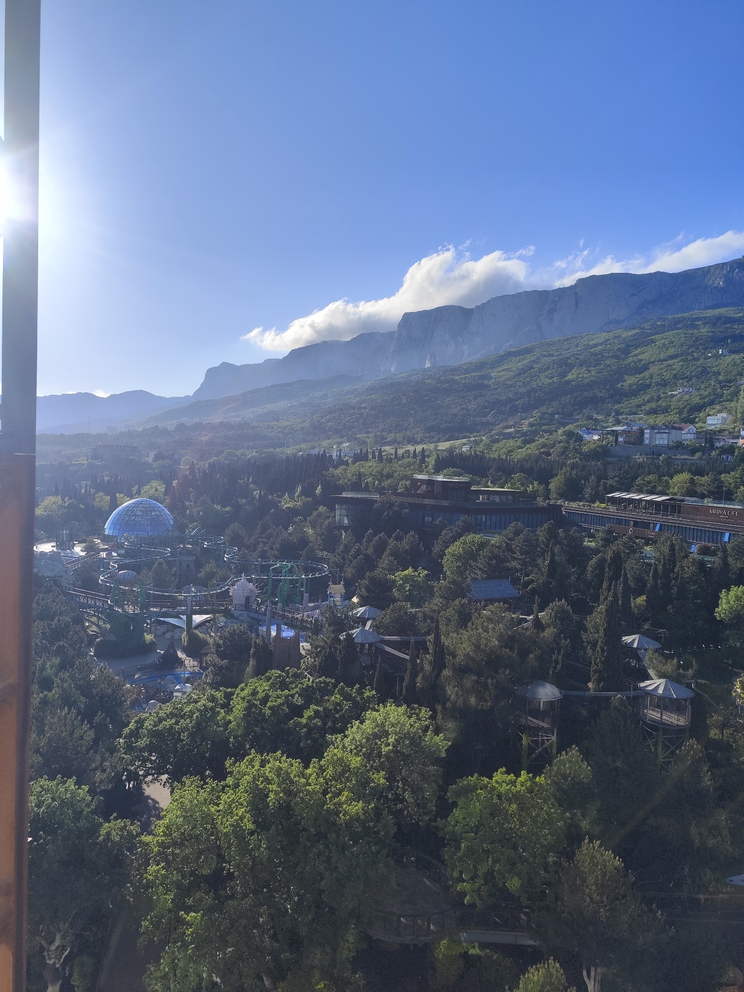 The image depicts a panoramic view of the city of Симеизский поселковый совет, Ukraine. In the foreground, there's dense greenery with trees. A blue geodesic dome structure is visible, likely part of an amusement park or entertainment complex with rollercoasters. Further back, there are buildings and hotels nestled within the landscape. In the background, there are prominent mountains under a clear blue sky with some clouds. The overall scene showcases a blend of urban development and natural landscape.