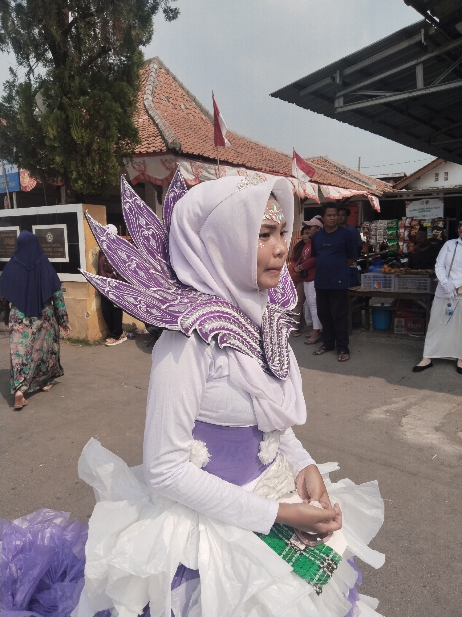 In Margasari, Indonesia, a young woman wearing a white hijab and a creative costume featuring large purple and white iridescent wing-like decorations around her neck is the central subject. The costume also includes a layered skirt made of white and purple plastic sheeting, and a purple sash around her waist, with a green plaid fabric detail visible at the hem. She has glitter applied to her forehead and cheekbones. The woman is positioned in the foreground, looking to her right.
In the background, a paved area is visible with several other individuals standing and walking. To the left, a woman in a dark blue hijab and a long floral dress walks away from the camera. Behind the main subject, several people are gathered, including men and women. One man in a blue t-shirt and black pants stands prominently. In the mid-ground, there are buildings with orange tiled roofs and awnings, suggesting a marketplace or street scene. Indonesian flags are visible on poles attached to one of the buildings. To the right, there are stalls or shops displaying various goods, including snacks and produce. The environment appears to be an outdoor, possibly public, space under a bright, overcast sky.