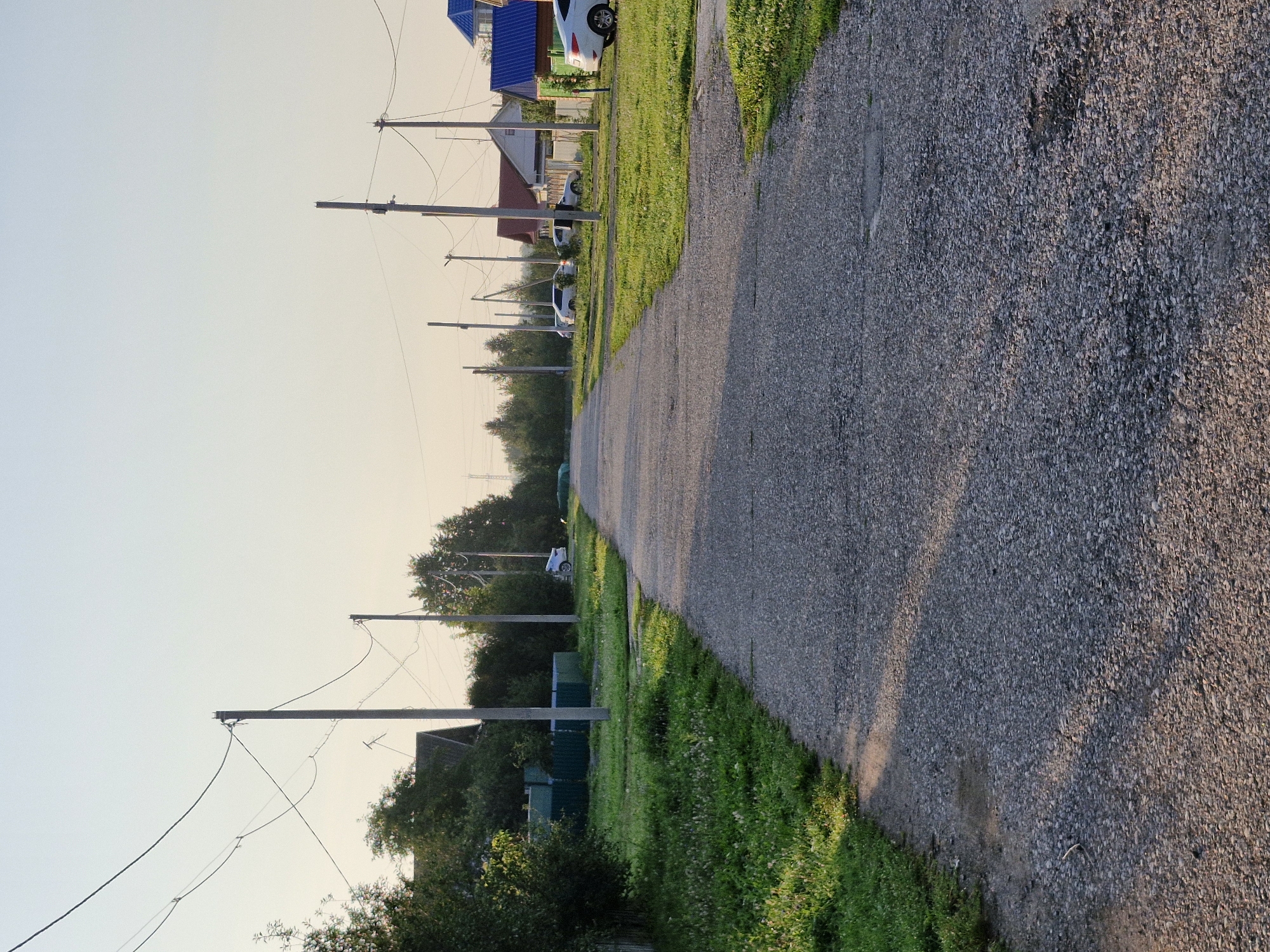 A two-lane asphalt road stretches into the distance, flanked by a grassy verge and residential buildings. Power lines run overhead, supported by wooden poles. Several white cars are parked along the roadside. The sky is clear and pale blue, suggesting daytime. The scene appears to be a quiet residential street in Buzdyak, Russia.