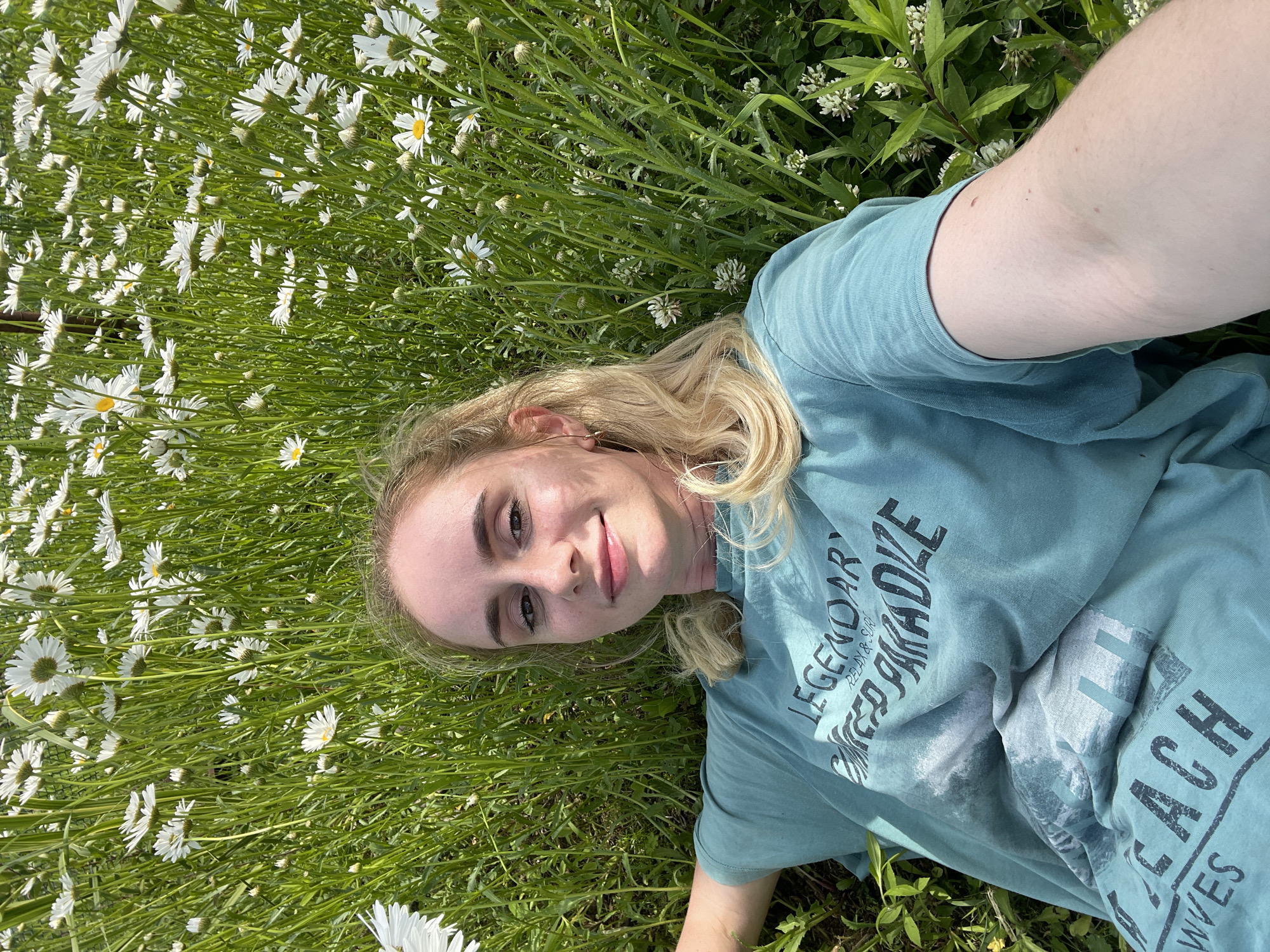 In Yasnoe, Russia, a young woman with blonde hair, wearing a teal t-shirt, lies on her back in a field of daisies and tall green grass, facing the camera. Her head is tilted slightly, and she is smiling. The t-shirt has a graphic print with text that reads "LEGENDARY," "RELAX & SURF," "PARADISE," and "BEACH WAVES." Her right arm is extended towards the camera, capturing a selfie. The background is filled with a dense meadow of white daisies and green foliage.