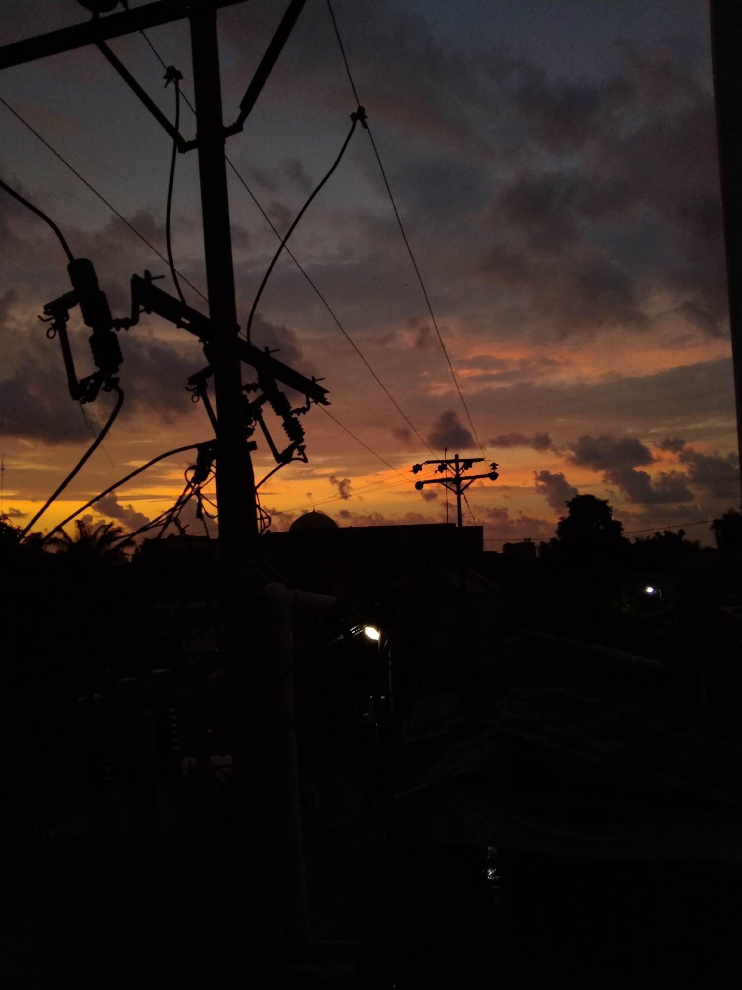 The image captures a serene sunset or dusk scene over what appears to be an urban or semi-urban area in Bireuenmeunasahdayah, Indonesia. The sky is a prominent feature, displaying a striking blend of deep orange, pink, and muted grey clouds, suggesting the transition from day to night. Silhouetted against this colorful backdrop are several utility poles and a complex web of power lines, dominating the left and central parts of the frame. One large utility pole stands prominently in the foreground, with its intricate structure and wires sharply defined against the lighter sky.
In the mid-ground, a distinct dome-shaped roof of a building, likely a mosque, is visible as a dark silhouette, further reinforcing the Indonesian context. Nearby, silhouettes of tropical trees, including what looks like palm trees, add to the local character. Another utility pole with crossarms is seen in the distance on the right, also silhouetted. Artificial light sources are scattered in the darker lower half of the image; a bright street light glows on the right side of the mid-ground, and a fainter light is visible on the far left. The foreground is largely dark and indistinct, showing the tops of buildings or rooftops. No people or visible text are present. The overall mood is quiet and atmospheric, highlighting the beauty of the fading daylight over a populated area.