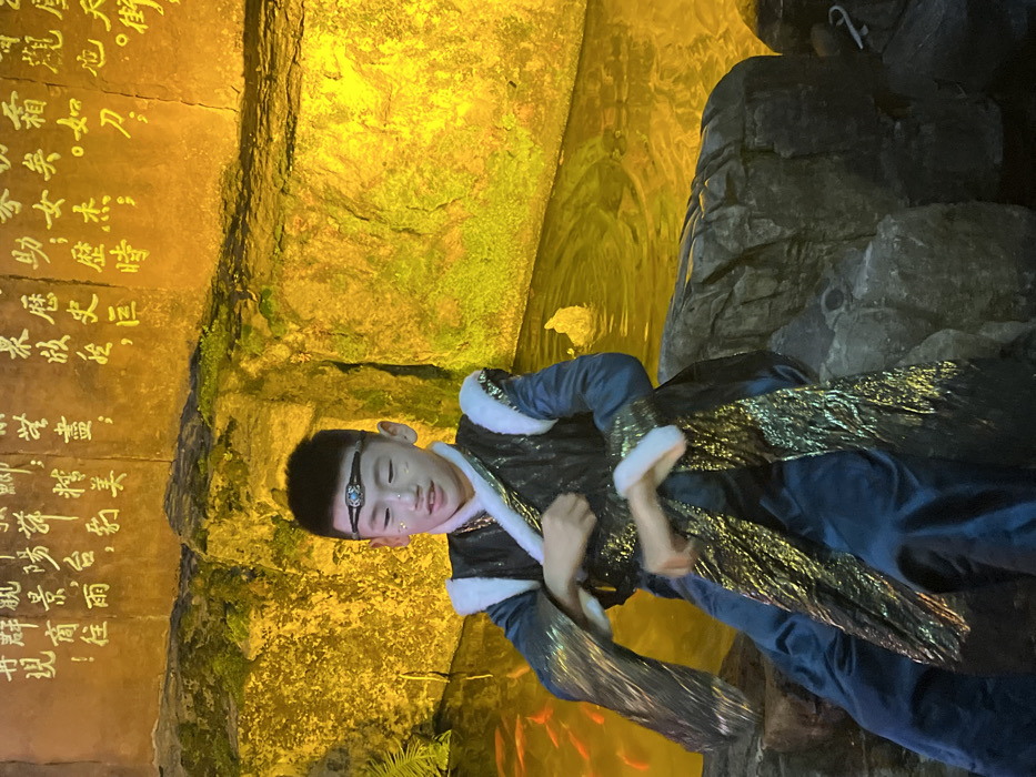 The image captures a young male in ceremonial attire seated on dark, rugged rocks beside a water feature, against an illuminated wall inscribed with Chinese characters, within Yuzhong District, China. The subject, a child or pre-teen with dark hair, wears a dark blue robe with white faux fur trim on the collar and cuffs, and an overlay of shimmering gold and dark textured fabric. His face features small, reflective glitter accents on the cheeks, and a dark headband with a central blue-green ornamental gem. His hands are raised and slightly blurred, indicating movement, possibly part of a performance or gesture. The background wall is bathed in a bright, golden-yellow light, highlighting the calligraphic inscriptions. The water feature reflects this golden light and contains several visible orange-red fish on the left, with a minor bubbling disturbance on the right.