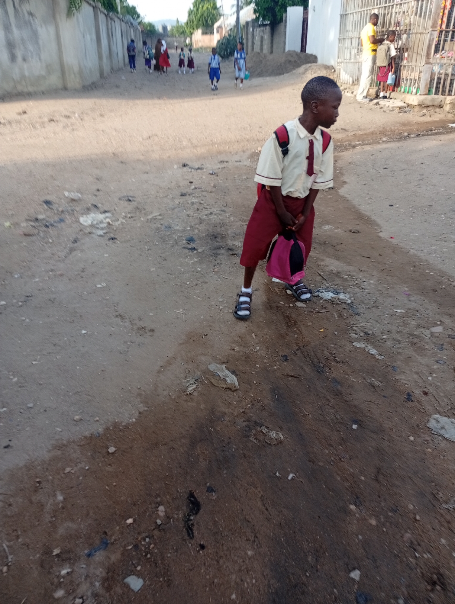 A young student stands in a dirt road in Adankolo, Nigeria. The student is wearing a school uniform of a tan shirt, red shorts, and black sandals, and carries a pink bag. Several other students are further down the road, walking away. A building with a barred window has people in front of it in the background. The road is dirt, with some debris and an area that appears wet. A concrete wall runs along one side of the road.