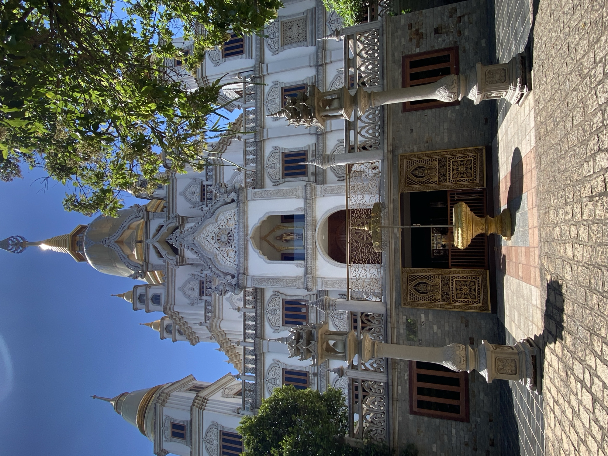 A detailed, slightly tilted, medium shot captures a portion of the Trúc Lâm Zen Monastery in Dĩ An, Vietnam. The monastery features a predominantly white facade, adorned with intricate ornamental detailing. The top of golden spires are visible against a clear blue sky. Parts of trees with green foliage are visible on either side of the building. The architectural style is characterized by layered structures, arched windows with dark blue frames, and elaborate stone carvings. Two ornate stone columns frame the entryway, leading to a golden gate with geometric floral patterns. The ground is paved with light-colored brick.