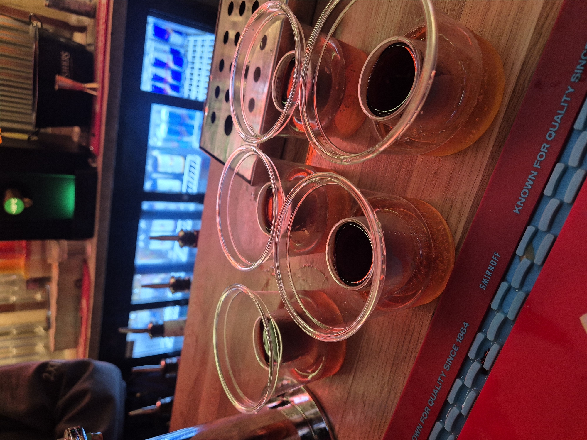 The image depicts a bar setting, possibly in West Suffolk, United Kingdom. Visible are six clear, rounded shot glasses filled with a liquid that appears to be layered with a dark top and a lighter amber bottom, suggesting a cocktail or mixed drink. These glasses are arranged on a wooden surface, likely the bar top. Adjacent to the glasses is a red object with the words "SMIRNOFF" and "KNOWN FOR QUALITY SINCE 1864" printed on it. In the background, a refrigerated display case containing beverage cans, potentially energy drinks, is visible. Behind the display case, assorted liquor bottles are discernible on shelves. A portion of a person's arm and sleeve can be seen in the bottom left corner.