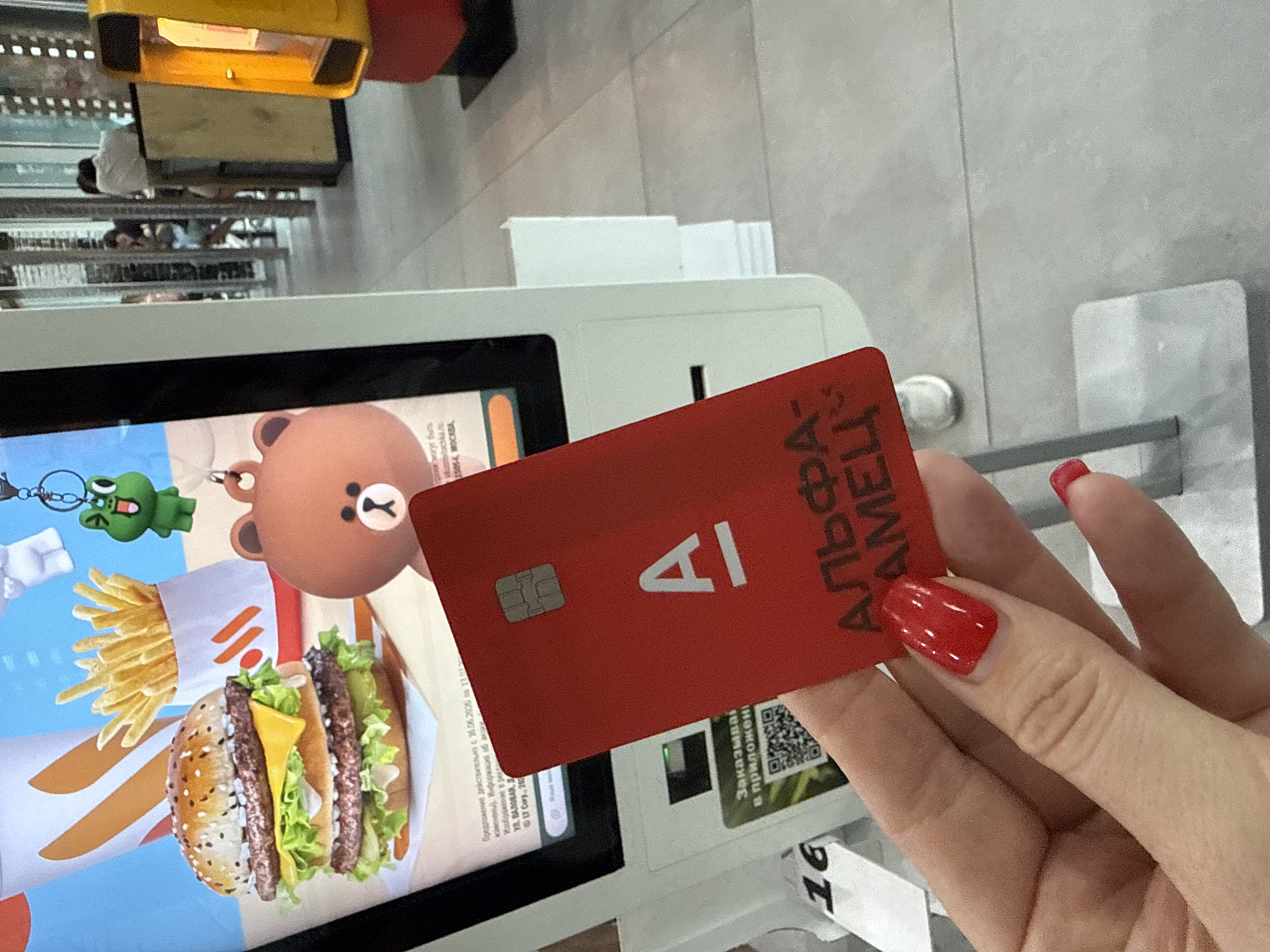 The image shows a close-up of a person's hand, with bright red nail polish, holding a red card with a silver chip and the logo "A" prominently displayed. The card also has text in Russian that appears to be an advertisement or offer from "ALFA-AMETS". The card is being held in front of a touch-screen kiosk, which displays an advertisement for a burger and fries, featuring cartoon characters. A QR code is visible on the kiosk screen. The background suggests an indoor commercial setting, possibly a fast-food restaurant or a similar establishment. The overall lighting indicates it is daytime, likely indoors as there are no external weather cues. The presence of a kiosk and advertisements suggests a transaction or ordering process is about to take place. The text on the card and kiosk is in Russian, and one of the location cues on the kiosk screen mentions "MOCKBA" (Moscow), although the provided location context is Sochi, Russia.