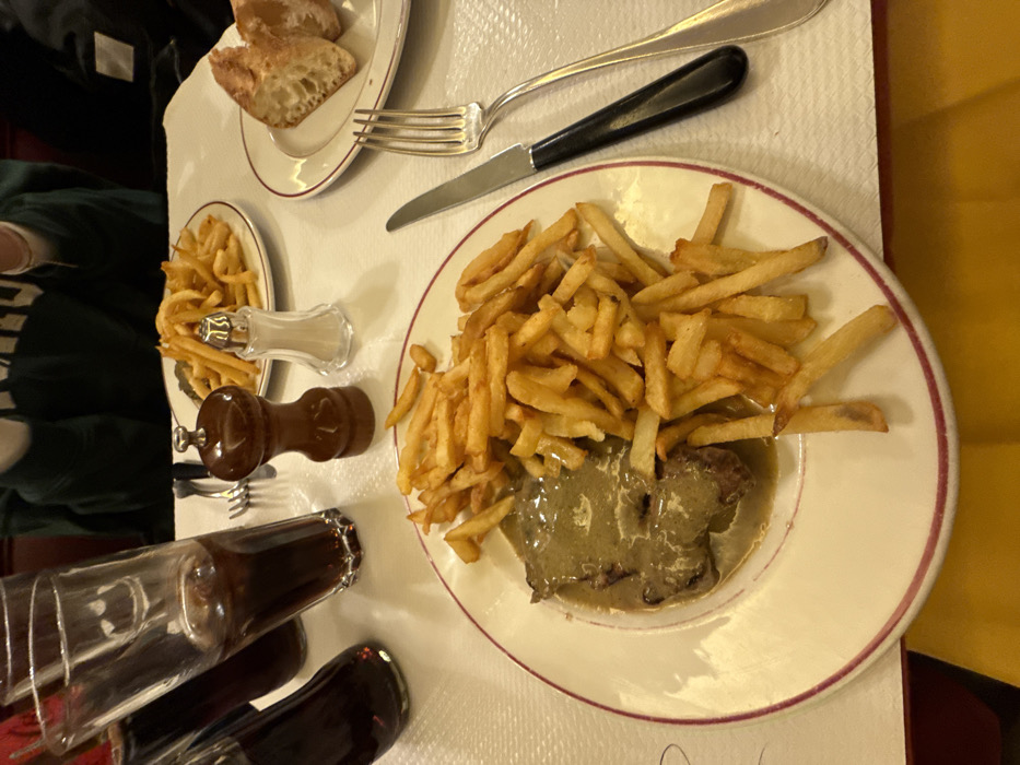 A plate of steak frites dominates the view, featuring a medium-rare steak covered in a creamy sauce, surrounded by a generous heap of golden-brown french fries. Salt and pepper shakers sit near a small side plate of more fries, suggesting a French restaurant setting. Nearby are two carafes of wine, along with a plate of bread, a fork and knife. The table is covered with a white tablecloth.