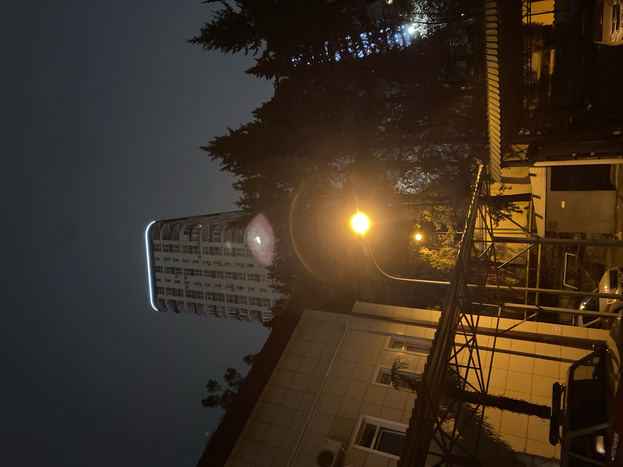 The image shows a street scene at night in Sochi, Russia. A tall, multi-story building with illuminated windows is visible in the distance on the left. A street lamp provides bright light. The right side of the frame is dominated by the facade of a lower building, likely a commercial or residential structure, and some scaffolding. A vehicle is partially visible, parked near the lower building. Some trees are visible in the background, and the sky is dark.