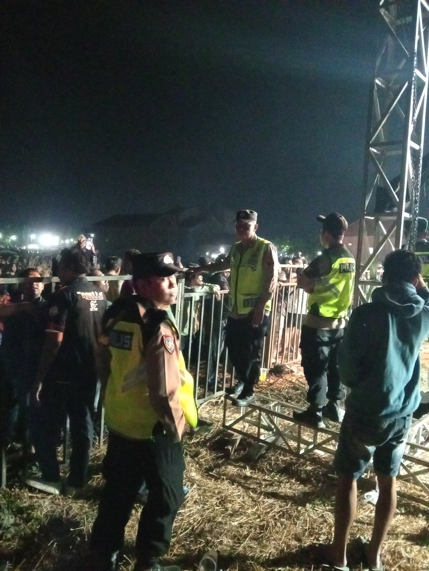 The image depicts a night scene at an outdoor event, likely a concert or festival, in Sukodono, Indonesia. In the foreground, three men in police uniforms are visible. The man on the left is wearing a black hat and a bright yellow vest with "POLISI" written on it. He is looking to his left. The man in the center is also in a yellow vest with "POLISI" and is facing away from the camera, with his arms crossed. The man on the right, partially obscured by the other two, is wearing a blue hooded sweatshirt and dark shorts.
Behind the police officers, a crowd of people is gathered, with some individuals holding up their phones, likely recording or taking pictures. A metal barricade separates the police from the crowd. In the background, a large metal structure, possibly a stage or lighting rig, is illuminated. The ground is covered with dry grass or straw. The overall atmosphere appears to be one of security and crowd control at a public gathering.