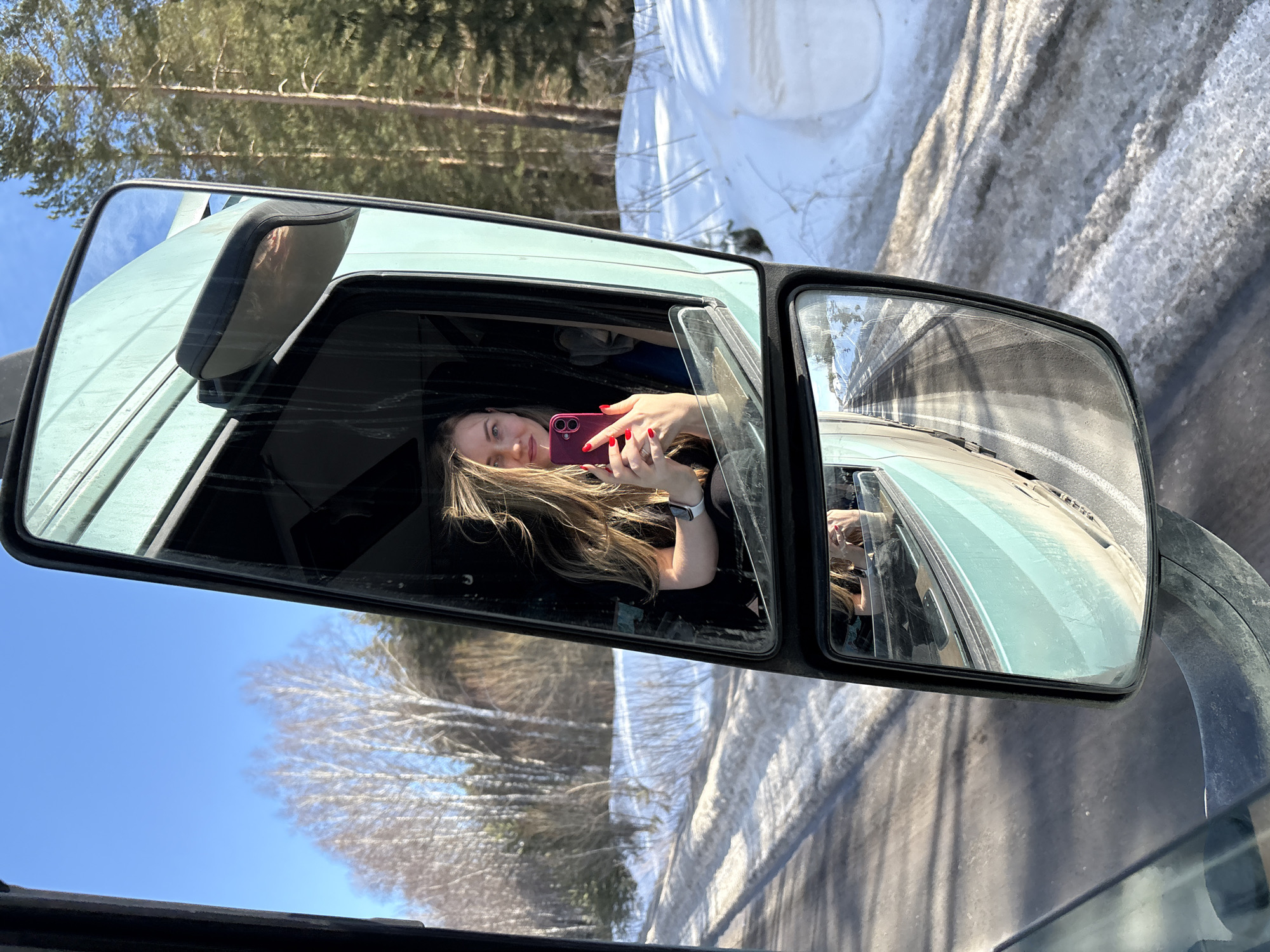The image displays a forensic view captured from inside a vehicle, looking towards its exterior side mirror in Iiul'skoe, Russia. The primary subject is a female reflected in the main rectangular side mirror. She has long, windblown light brown hair and is wearing a dark garment. Her left wrist bears a white smartwatch. She holds a magenta-colored smartphone with her right hand, displaying red-painted fingernails, and is smiling directly at the camera's perspective, indicating a selfie. The exterior of the vehicle, visible around the mirror, is a light green or teal color. Below the main mirror, a smaller convex mirror reflects the road, showing a dark asphalt surface with visible tire tracks and patches of melting snow and slush. The surrounding environment consists of a clear blue sky overhead. To the left of the vehicle, bare deciduous trees, possibly birches, stand against the sky. To the right, a dense coniferous forest, likely pine, is visible. Significant snow accumulation lines the roadside and forms mounds, consistent with a winter or early spring setting. The road's linear perspective in the lower mirror suggests the vehicle is in motion.