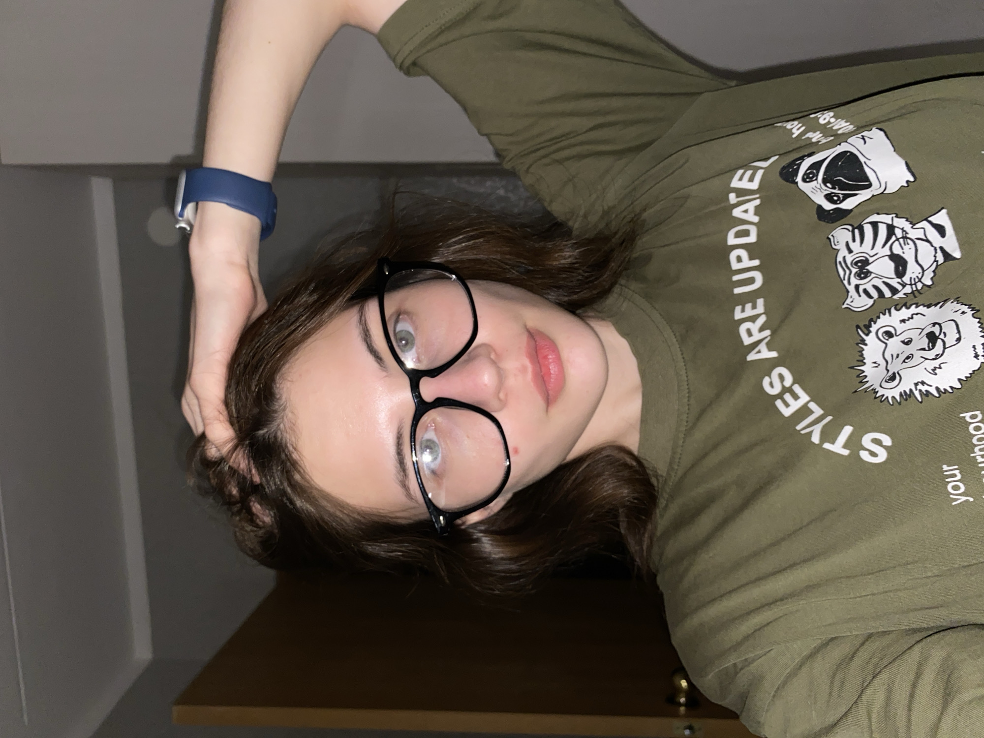 A young woman is posing for a selfie indoors. She is wearing glasses with black frames and a green t-shirt that features cartoon animal drawings and the words "STYLES ARE UPDATED your neighborhood." She has brown hair and fair skin. Her left arm is raised, with her hand touching her hair. A blue smartwatch is visible on her left wrist. The background consists of a partially visible shelf, a white wall, and a dark wall. There is no information available to determine if this image was taken in Votkinsk, Russia.