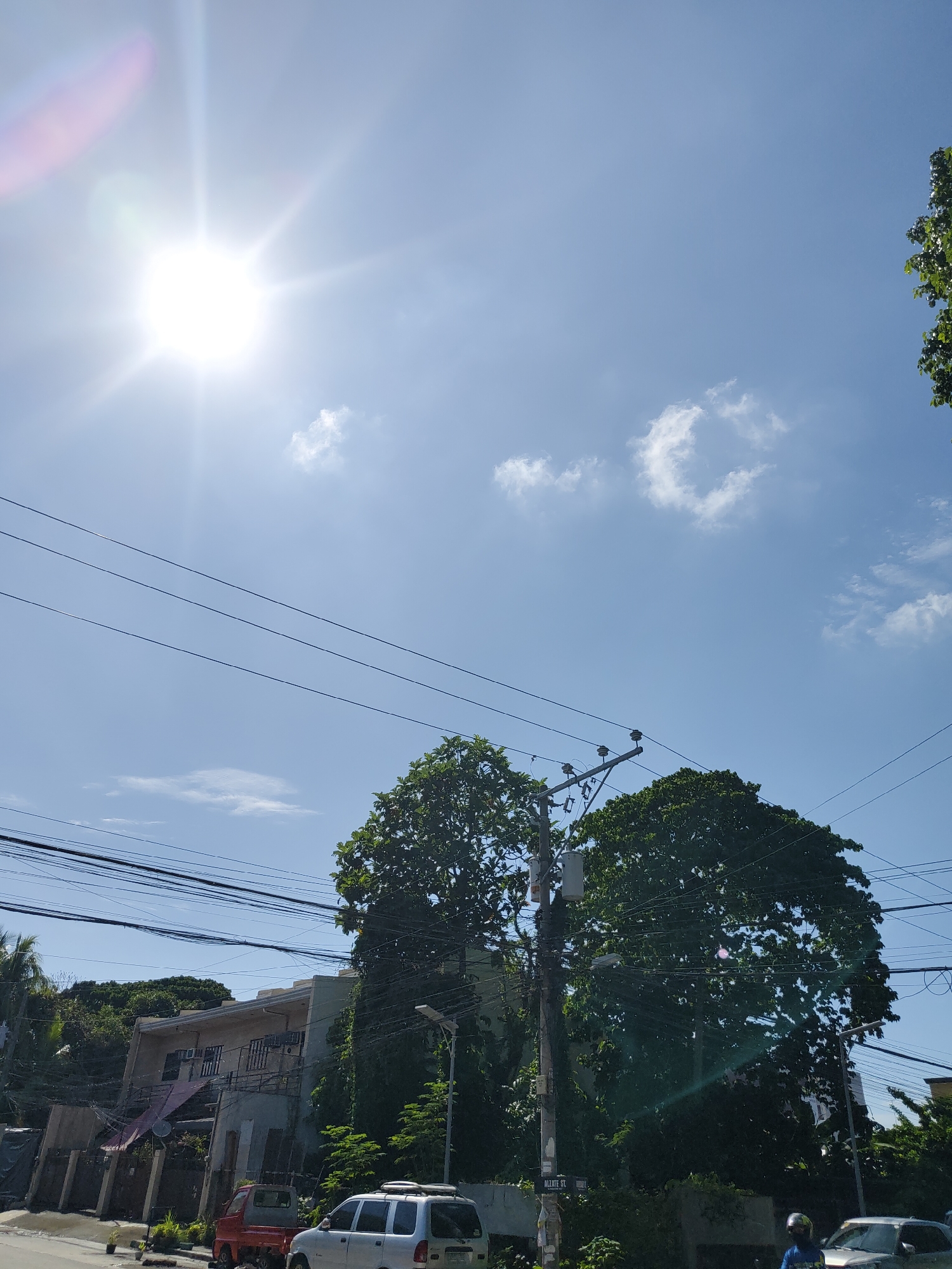 The image depicts a vibrant, sunlit street scene, likely captured in Davao City, Philippines, given the location context. The upper portion of the frame is dominated by a clear, bright blue sky with a few scattered white clouds. The sun shines intensely from the upper left, producing prominent rays and lens flare.
A dense web of overhead power lines and communication cables traverses the sky, connecting to several utility poles. One significant utility pole stands in the mid-ground, heavily laden with electrical components and wires. Another pole in the lower right corner bears a street sign.
Lush, green trees with abundant foliage populate the mid-ground and foreground, partially obscuring the background structures. Behind the trees and wires, a multi-story building with multiple windows, balconies, and air conditioning units is visible, suggesting a residential or mixed-use property.
On the paved street in the lower part of the image, a small red pickup truck is parked on the left, and a silver SUV or van is parked on the right. A person wearing a blue shirt and a helmet is partially visible on the far right, near another vehicle, possibly a motorcycle.
The scene conveys a bright, clear day, with the high position and intensity of the sun indicating it is likely mid-day. The only visible text is "ALLATE ST." on a street sign affixed to a utility pole.