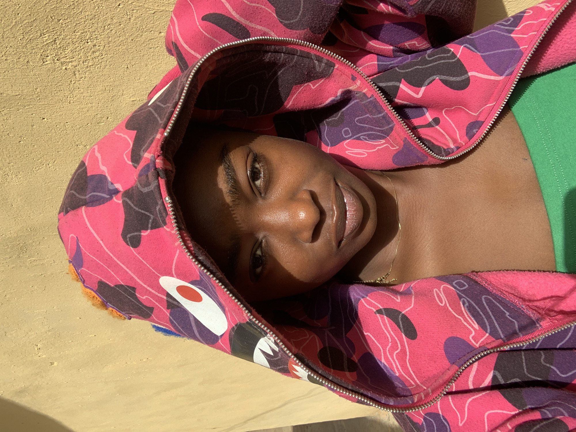 A person with dark skin, wearing a pink and purple camouflage hoodie, lies in a sunlit area. The person's head is within the hoodie, and the hood is zipped up but open, partially revealing the person's face. The hoodie has a distinctive design featuring a large white eye and red pupil. A green garment is also visible beneath the hoodie. The person is wearing a gold necklace. The background is a textured, beige surface. This image was taken in Rafin Par, Nigeria.