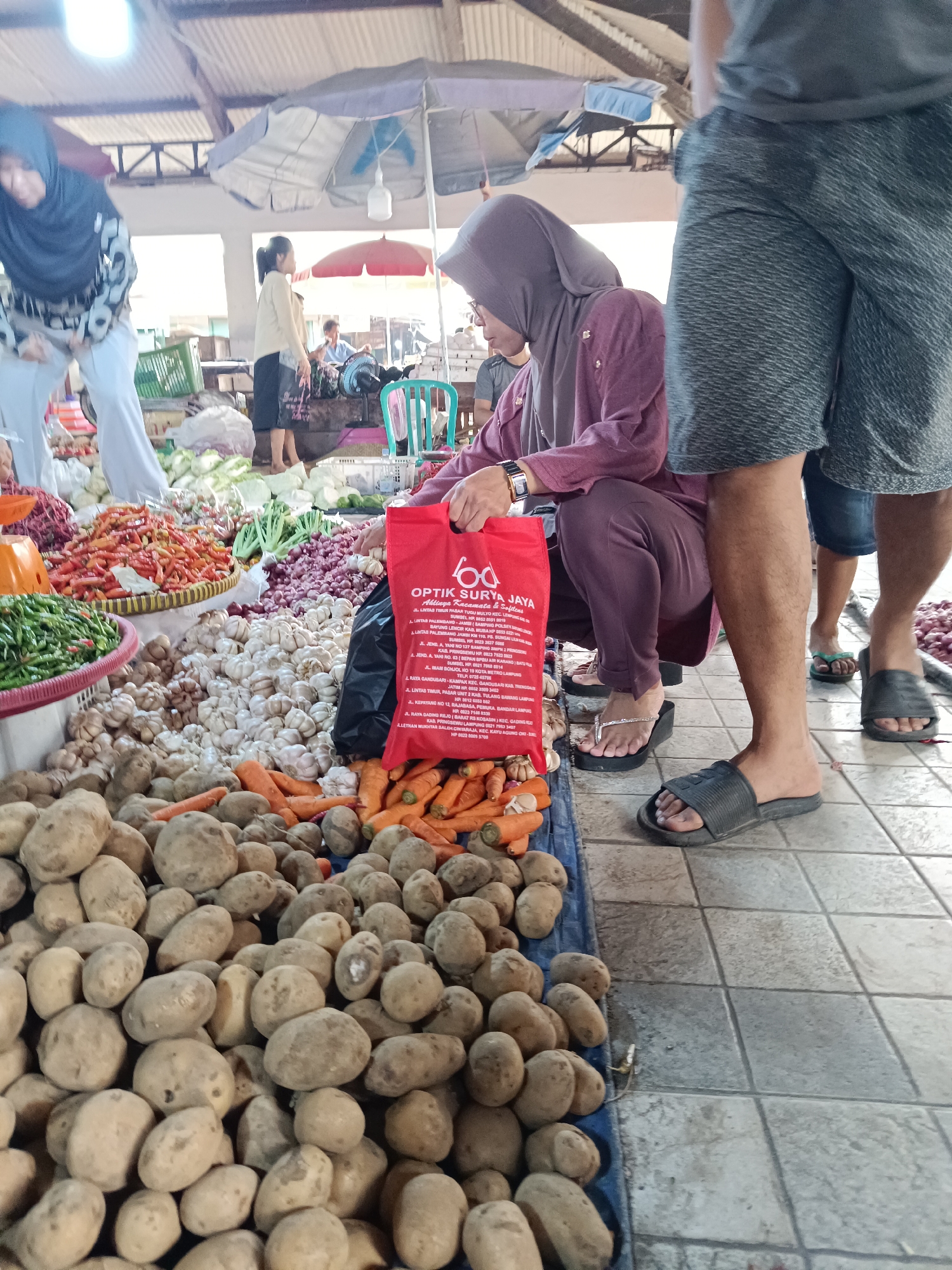 The image depicts a bustling open-air market in Kayu Agung, Indonesia, likely focused on fresh produce. In the foreground, a large pile of potatoes dominates the bottom left of the frame, with carrots and garlic bulbs scattered amongst them. To the right, a person wearing a purple hijab and traditional clothing is squatting down, holding a red reusable shopping bag with "OPTIX SURYA JAYA" printed on it in white text. Another person's legs and feet, clad in grey shorts and black flip-flops, are visible in the right foreground, suggesting they are standing. In the background, more produce, including red chilies and green onions, is displayed, along with other market-goers and stalls. The market appears to be covered by a corrugated roof supported by metal beams, with natural light filtering in. Several umbrellas and tarps provide additional shade. The overall scene is one of vibrant commerce and everyday life in a local Indonesian market.