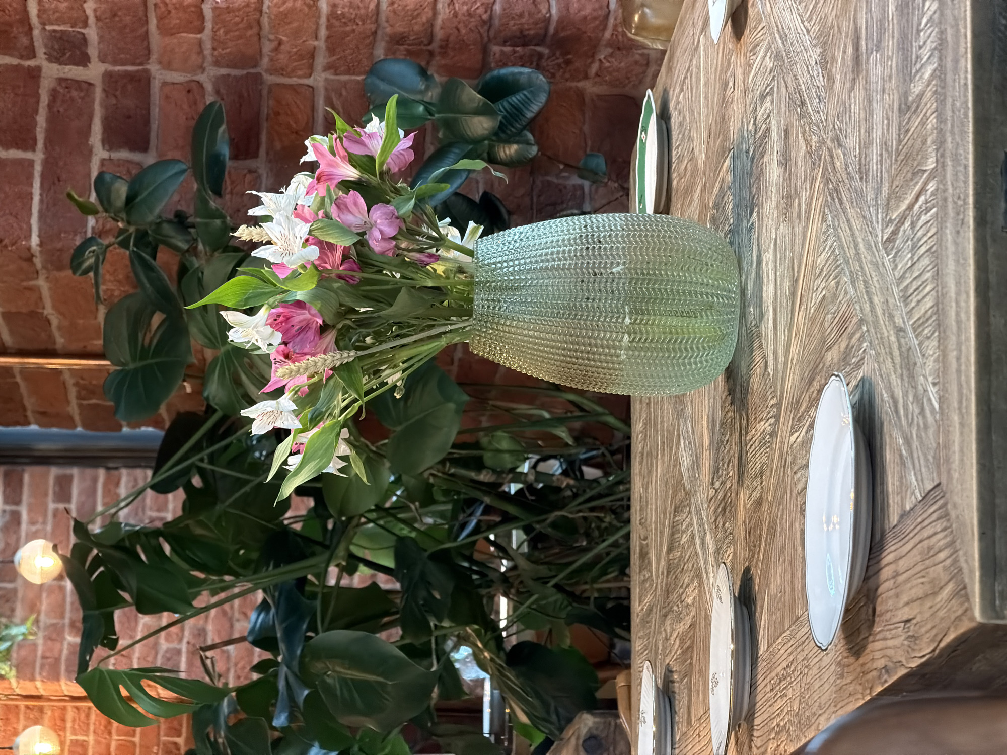 This media file captures a still life scene featuring a bouquet of pink and white alstroemeria flowers in a textured, light green glass vase. The vase sits on a rustic wooden table with a distinct chevron pattern. To the left of the vase, a large, leafy plant with dark green, broad leaves is visible, its branches reaching towards the top of the frame. Behind the plant and vase, a brick wall with a warm, reddish-brown hue serves as the backdrop. The lighting suggests an indoor setting, possibly with artificial light sources, as evidenced by two glowing orbs that appear to be lights on the lower left side of the image. Several white plates with delicate gold trim are arranged on the wooden table, with the largest plate prominently positioned to the right of the vase. The overall impression is one of a well-decorated, perhaps domestic or restaurant setting. The lighting and composition create a somewhat intimate and serene atmosphere. There is no visible text in the image. The location context of Saint Petersburg, Russia, does not provide any further visual cues within the image itself.