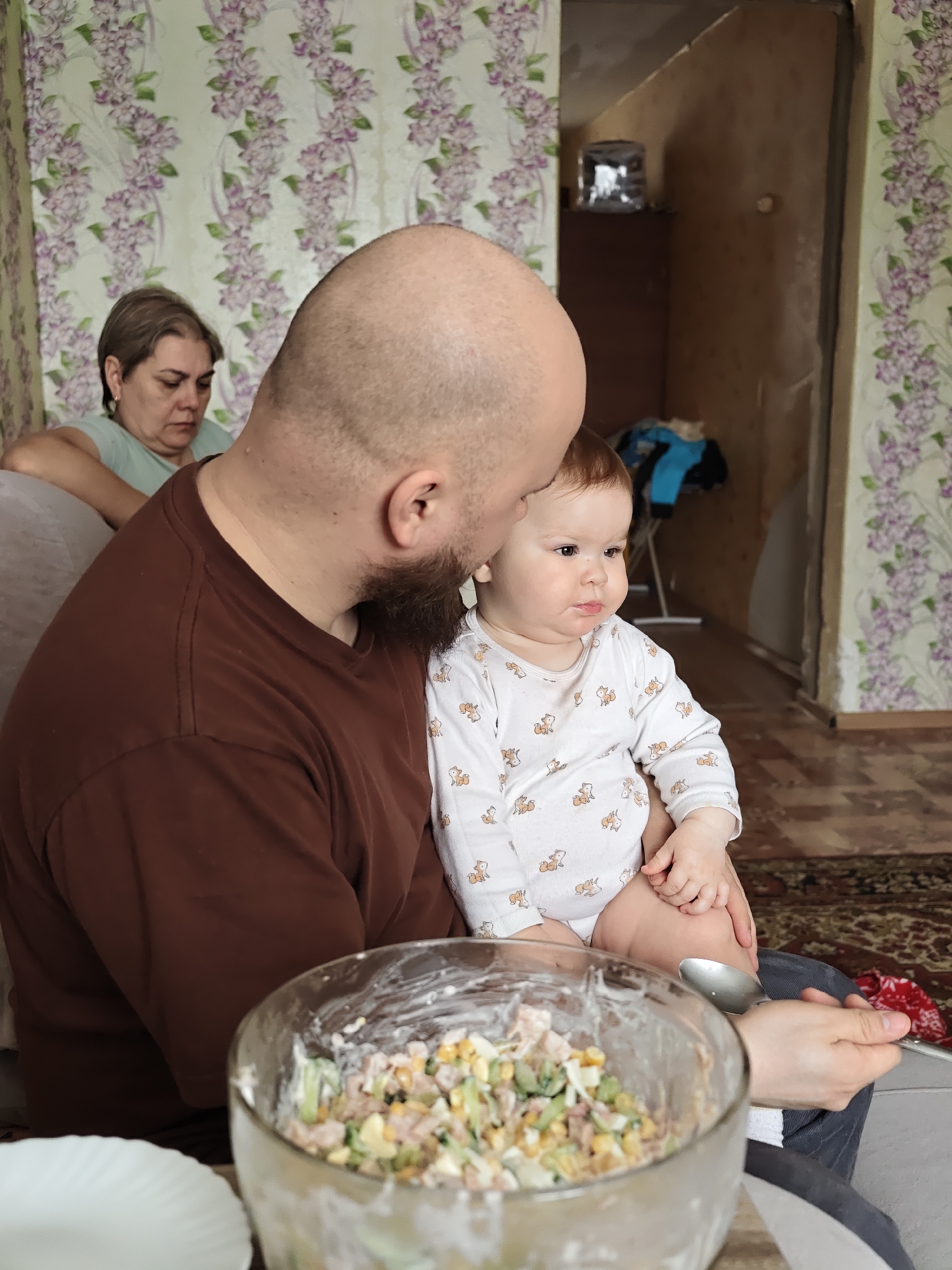 Here is a forensic description of the image:
The image, apparently taken in Уральское, Russia, shows an indoor scene. A bald man with a beard, wearing a brown t-shirt, is holding a baby wearing a white onesie with orange dog prints. The baby is looking off to the right side of the frame. In front of the man is a glass bowl containing salad. A spoon is visible near the bowl. A white plate sits to the side. Behind the man and to the left is a woman with short hair, sitting on a couch. The wallpaper has a floral pattern in green and purple. A doorway leads to another room where clothing is visible on a rack. The floor appears to be carpeted.