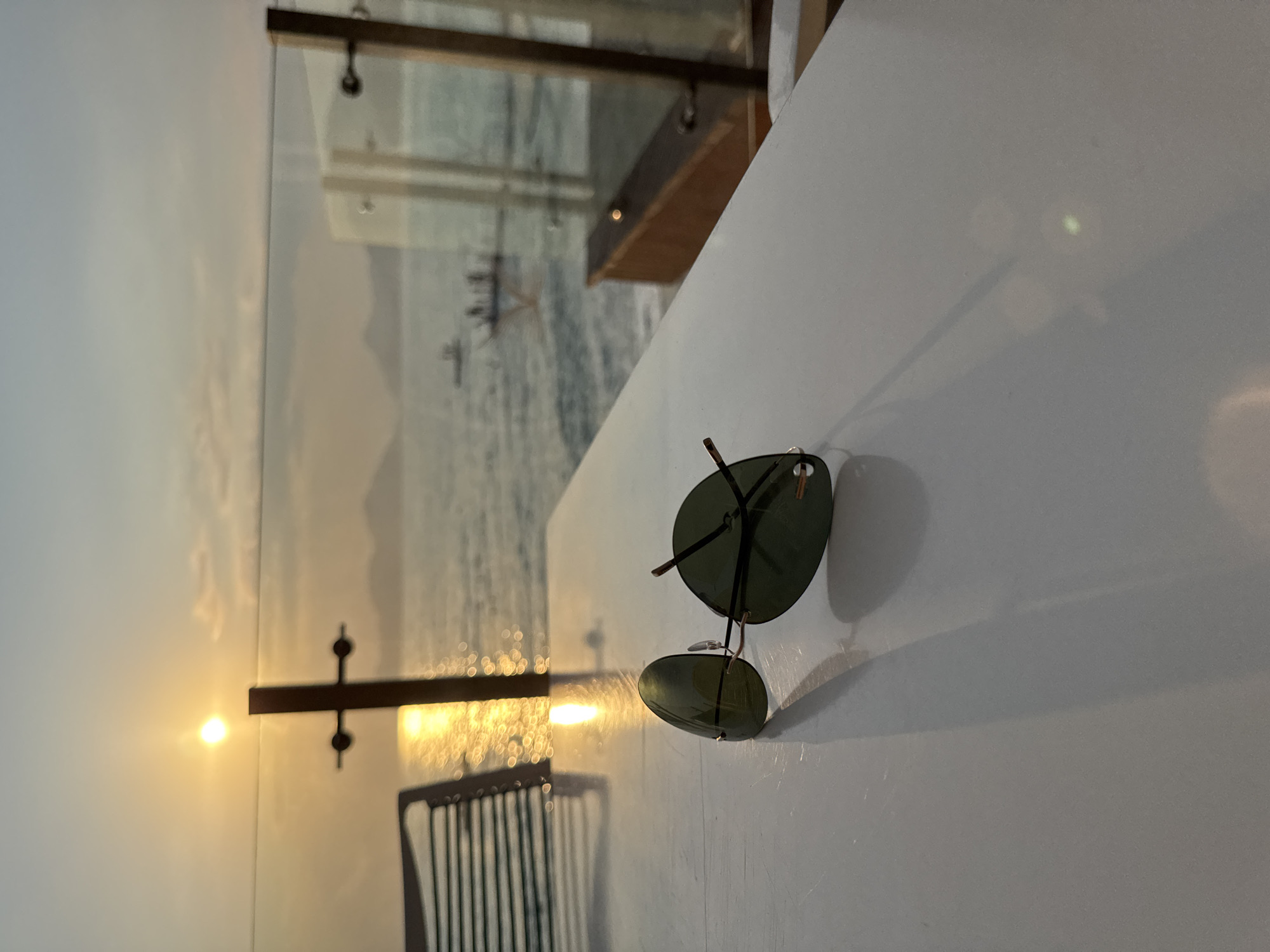 A pair of aviator sunglasses rests on a white table with a blurred background of the sea at sunset. The sun is visible in the sky, casting a golden reflection on the water. In the distance, silhouettes of boats and mountains can be seen. The scene appears to be a serene outdoor dining area or balcony overlooking the sea. It is likely located in Fethiye, Turkey, a popular tourist destination known for its beautiful coastal scenery and sunsets.