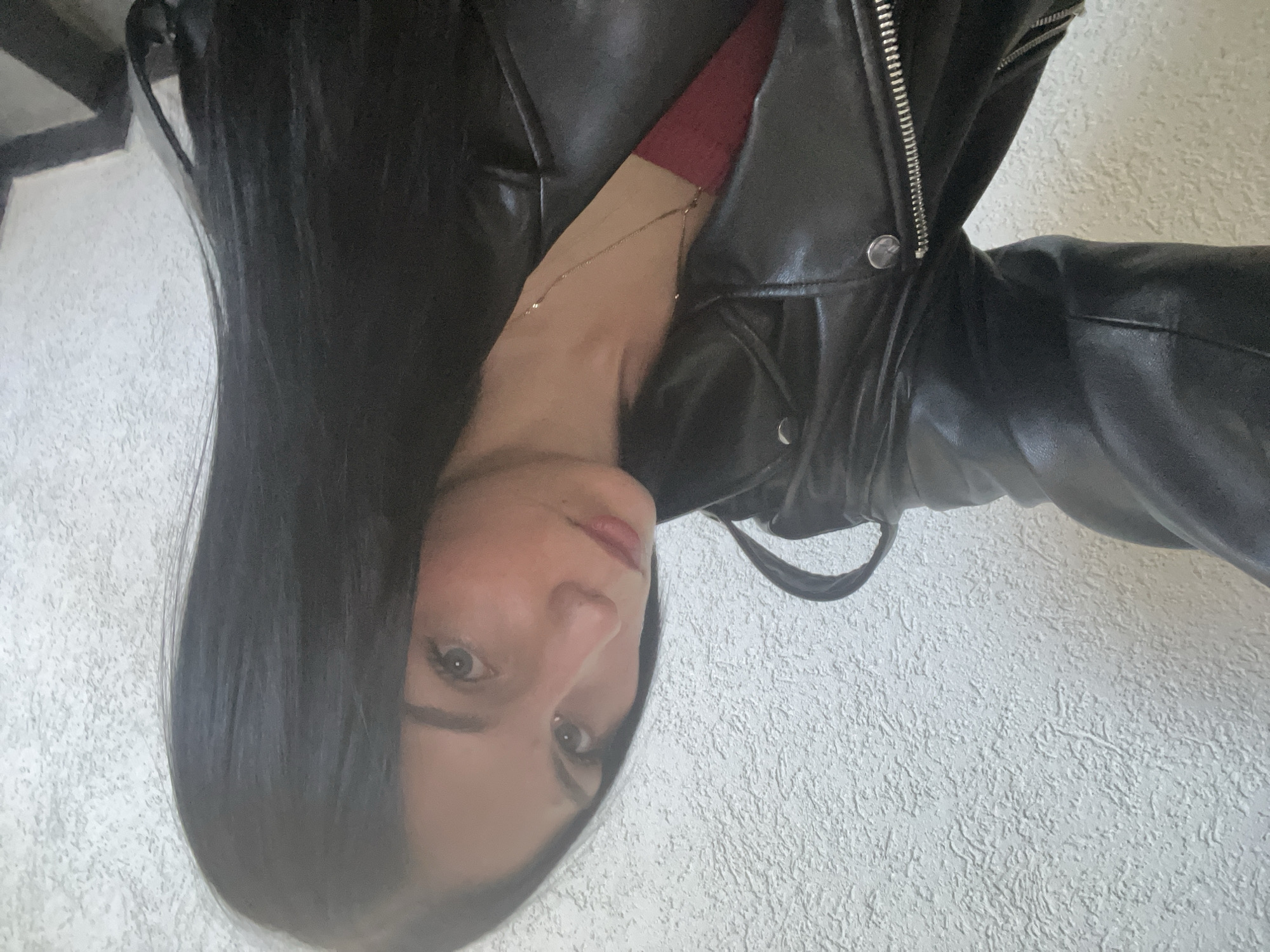 A young woman with long, dark hair and fair skin poses for a selfie against a textured, off-white wall. She is wearing a black leather jacket over a dark red top, and a delicate gold necklace with small beads. The image captures her from the chest up, with her face turned slightly towards the camera. The background is a plain wall, and to the right, a dark staircase with black steps and risers is visible.