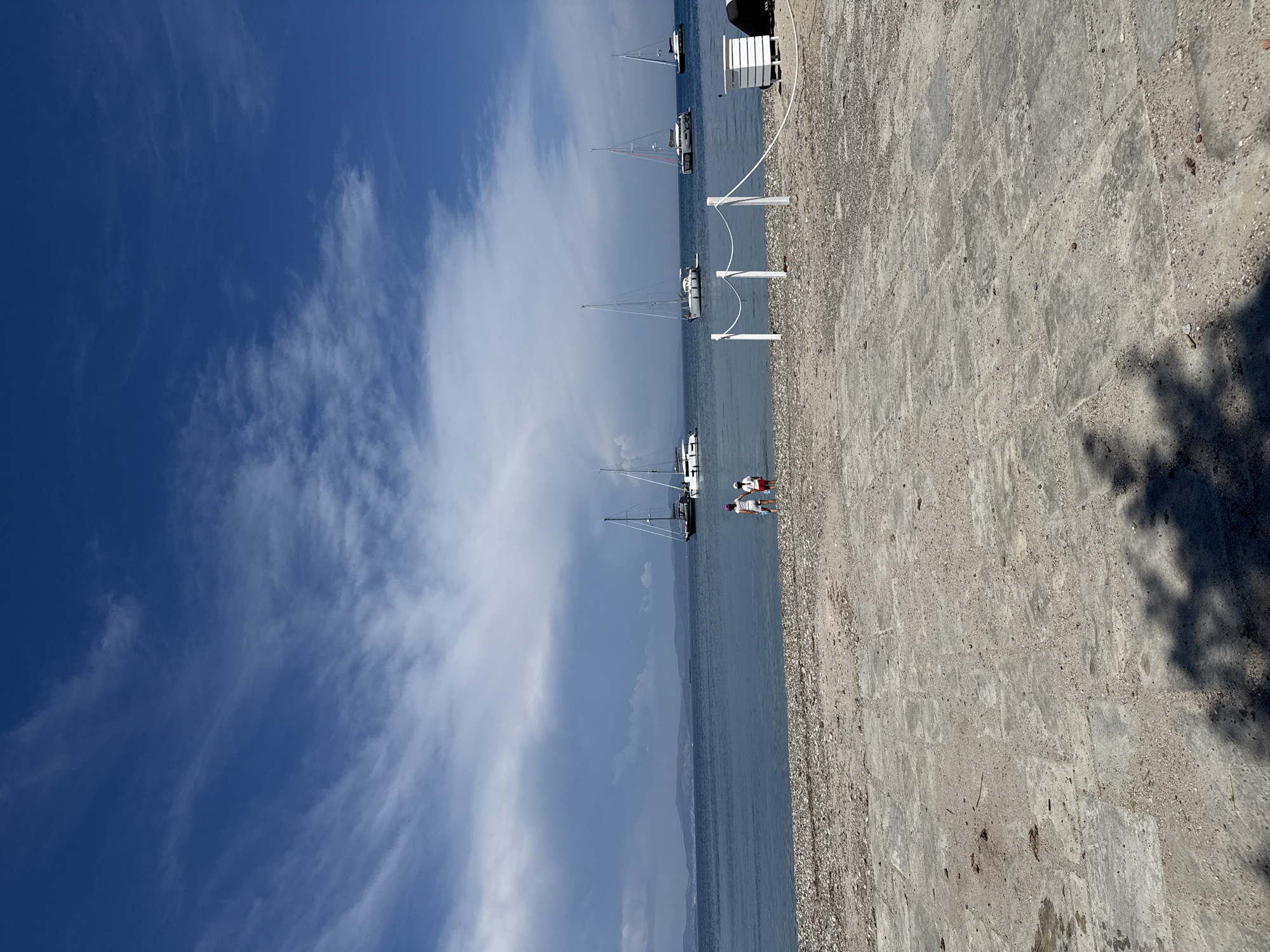 This image depicts a waterside scene in Kos, Greece. The composition is dominated by a clear blue sky with scattered white clouds to the left, and a line of sailboats anchored offshore along the center. The foreground features a textured stone or concrete walkway. Two people are seen standing near one of the boats. White bollards or posts, possibly for mooring boats, run along the edge of the walkway. The overall scene suggests a sunny day at a coastal location in Kos, Greece.