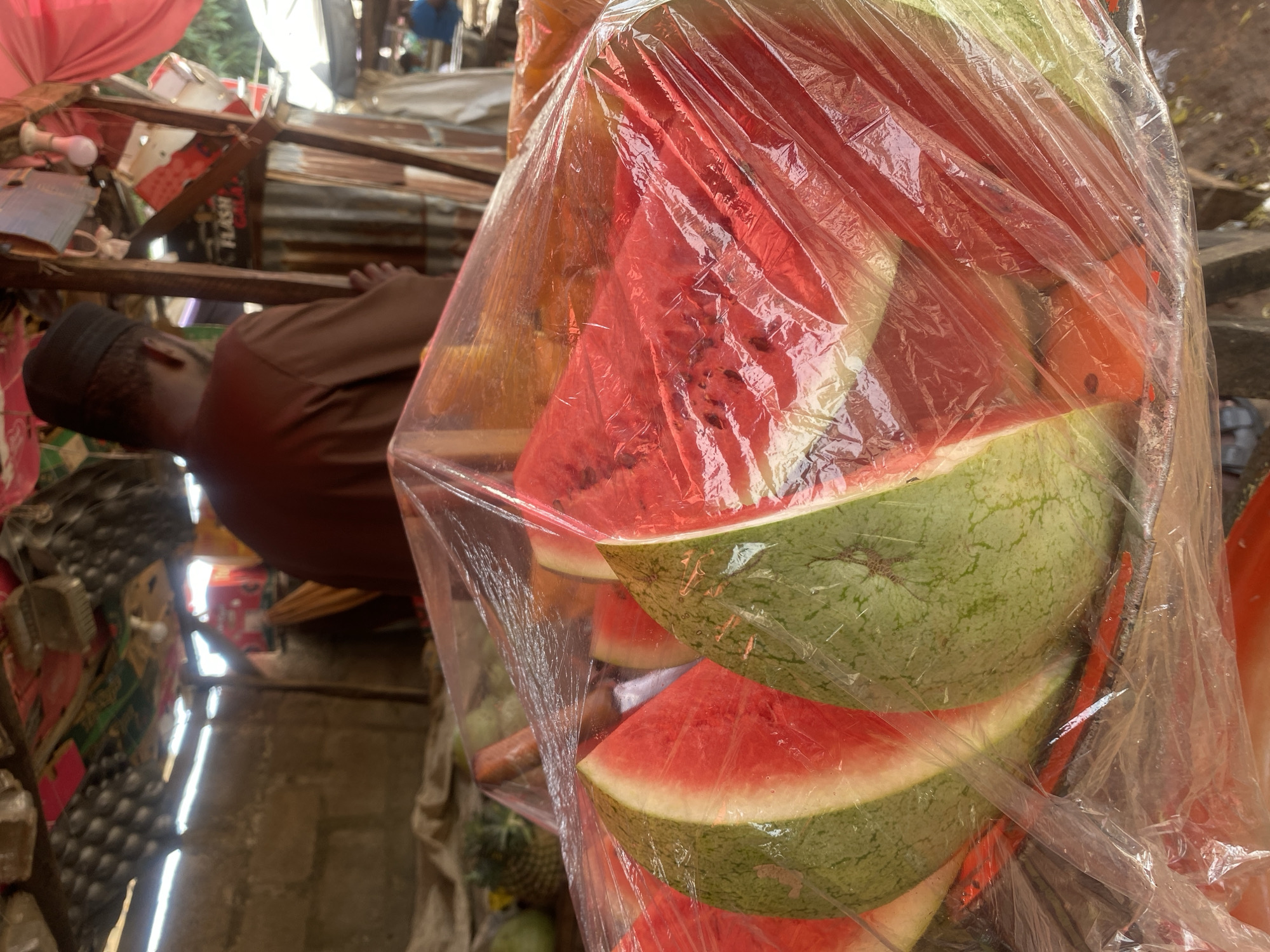 The image depicts a fruit stall located in a market setting within Sabon Tasha, Nigeria. In the immediate foreground, several large, triangular slices of red watermelon with green rinds are prominently displayed on a tray and enclosed in clear plastic wrap. Behind these watermelons, a person, identified as male, is visible from the back, wearing a brown tunic and a dark cap. He stands within a rustic market stall constructed from corrugated metal sheets, rough wooden beams, and an overhead canopy composed of mixed materials, possibly including cardboard and fabric. Various other fruits, including pineapples and what appear to be carrots, are visible on a surface behind the main watermelon display. A partially obscured sign displaying the text "FLASH GALA" is attached to a wooden beam on the right side of the frame, near an exposed incandescent light bulb. The environment suggests an active, semi-enclosed open-air market.