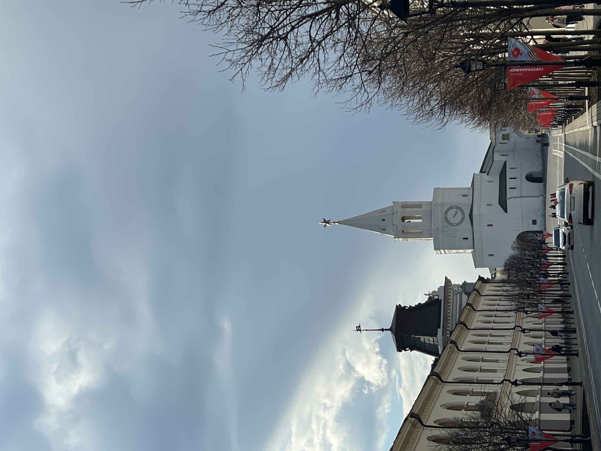 The image depicts an urban street scene in Kazan, Russia, under a bright, overcast sky. The photograph is oriented vertically, showing the sky at the bottom and the ground at the top of the frame in its original state; for descriptive purposes, it will be described as if rotated 90 degrees counter-clockwise to a standard horizontal view.
On the right side of the frame, the prominent white Spasskaya Tower of the Kazan Kremlin stands tall, featuring a visible clock face and a golden five-pointed star atop its spire. On the left, a historic-looking building with a beige facade, multiple arched windows, and a distinctive dark, ornate roof with a small decorative spire is visible.
A wide street or avenue runs through the middle ground, with visible road markings. Several vehicles, including a white van, are on the road, and pedestrians can be seen walking on the sidewalks along both sides of the street. Bare trees line the thoroughfare, suggesting the season is late winter or early spring.
Numerous red banners are attached to the street lamps. Visible text on these banners includes "ГОРЖУСЬ ТОБОЙ!" (I am proud of you!), "1945-2025," and "С ДНЁМ ПОБЕДЫ!" (Happy Victory Day!). These inscriptions indicate a public celebration or commemoration related to Victory Day, likely for its 80th anniversary in 2025. The overall setting is a significant urban area with historical architecture.