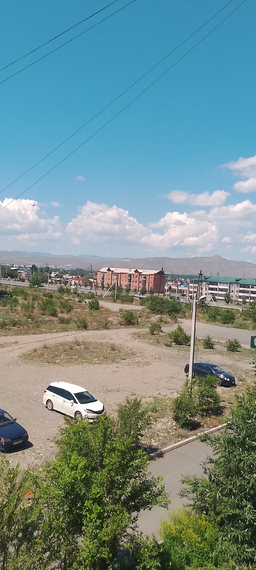This is a medium-shot, eye-level, outdoor, vertical photograph taken on a sunny day in the city of Kyzyl, Russia. The sky is bright blue with scattered white clouds. Several power lines are visible in the upper portion of the frame.
In the mid-ground, there is a large, multi-story brick building with a light-colored roof and balconies. To the right of this building are several other buildings, some with green roofs. Power poles and lines are present throughout this area. The terrain is mostly dry and dusty with sparse vegetation.
In the foreground, on a dirt and gravel surface, are three cars: a dark blue sedan on the left, a white station wagon in the center, and a dark gray sedan to the right. The white station wagon appears to be parked. Lush green trees are visible in the immediate foreground, obscuring the lower portion of the scene. The background shows rolling hills under a bright sky.