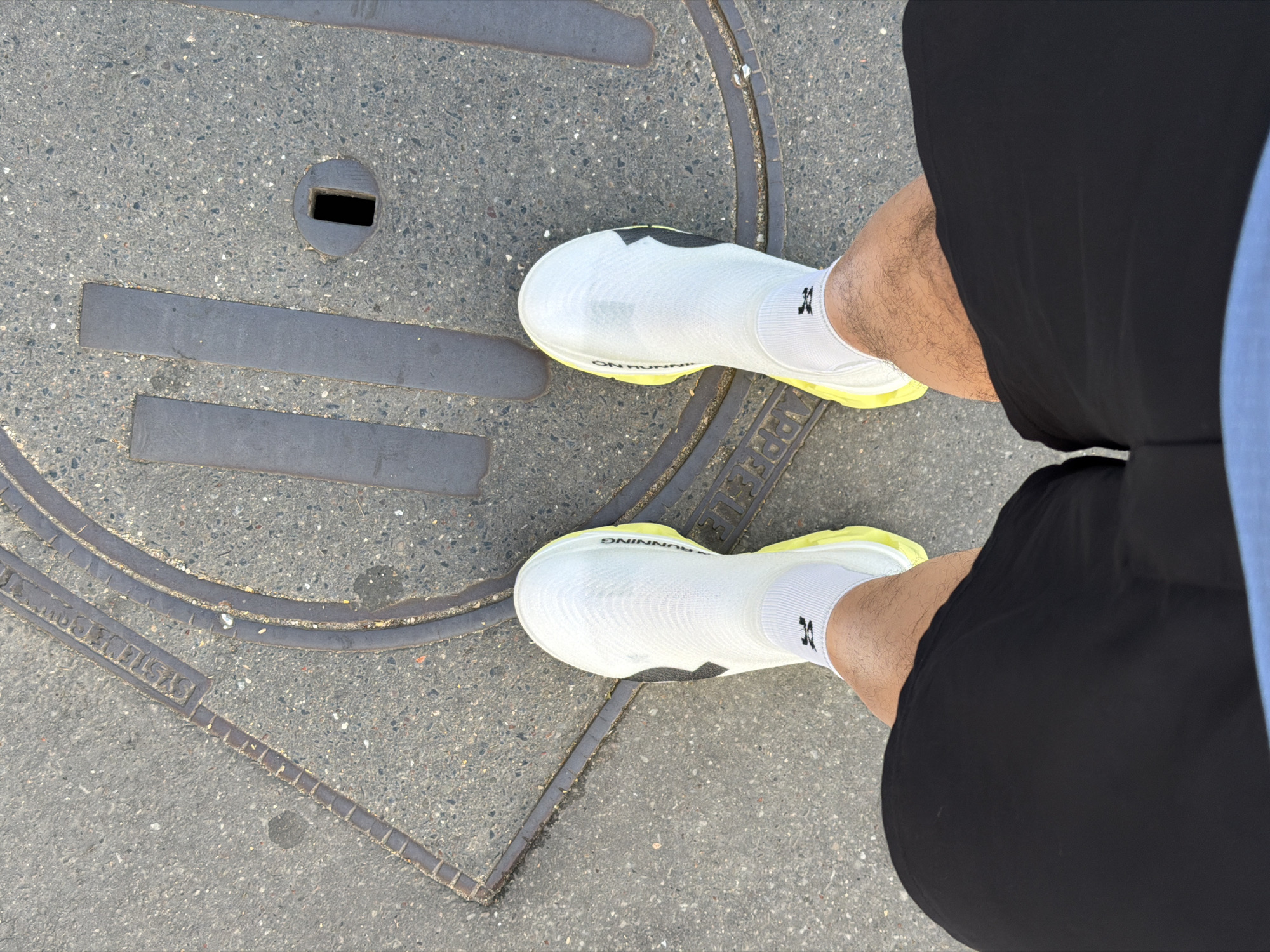 A person wearing black shorts, white running shoes, and white socks stands on a round manhole cover on a paved street. The manhole cover is set in concrete and has a series of raised, linear metal bars and concentric circles around its perimeter, as well as a rectangular inset. Part of the text "APPROFC" is visible on the cover. The location is consistent with urban pavement, which could be in the city of Paris, France.