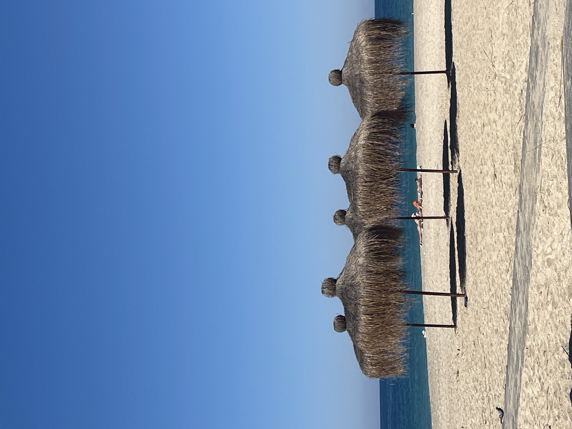 The image shows a beach scene in Alanya, Turkey, under a clear blue sky. Four straw parasols are lined up along the beach. The sand is light-colored and has variations in texture and patterns. The sea is visible in the background, with a transition in color from turquoise to a darker blue further out. Several people are lying on the beach under the parasols.
