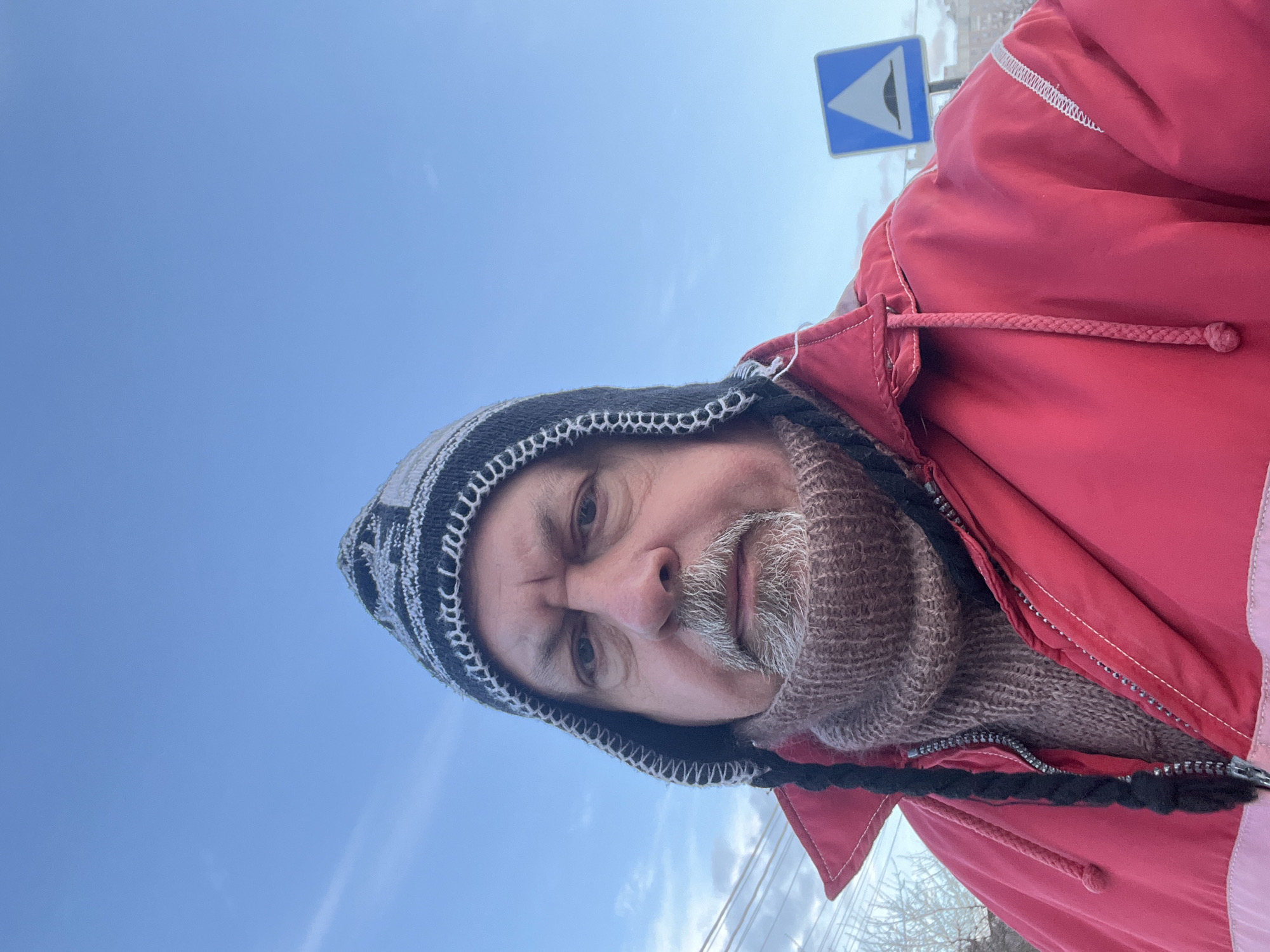 The image presents a close-up, outdoor portrait of a male individual, appearing middle-aged or older, positioned centrally in the frame. He has a grey beard and mustache and is looking directly at the camera. The subject is wearing a black and white patterned knitted winter hat with ear flaps and braided ties, a brown knitted turtleneck, and a partially unzipped red winter jacket. The jacket features a lighter pink horizontal stripe near its lower edge and white decorative stitching on the right shoulder area.
The background primarily consists of a clear blue sky with scattered thin white clouds. To the left of the subject, bare tree branches and utility lines are visible. To the right, a blue square road sign with a white upward-pointing triangle and a black speed bump symbol is prominently displayed. Faint outlines of light-colored buildings are discernible in the distant background, partially obscured by the sign. The scene is situated outdoors in the city of Podolsk, Russia.